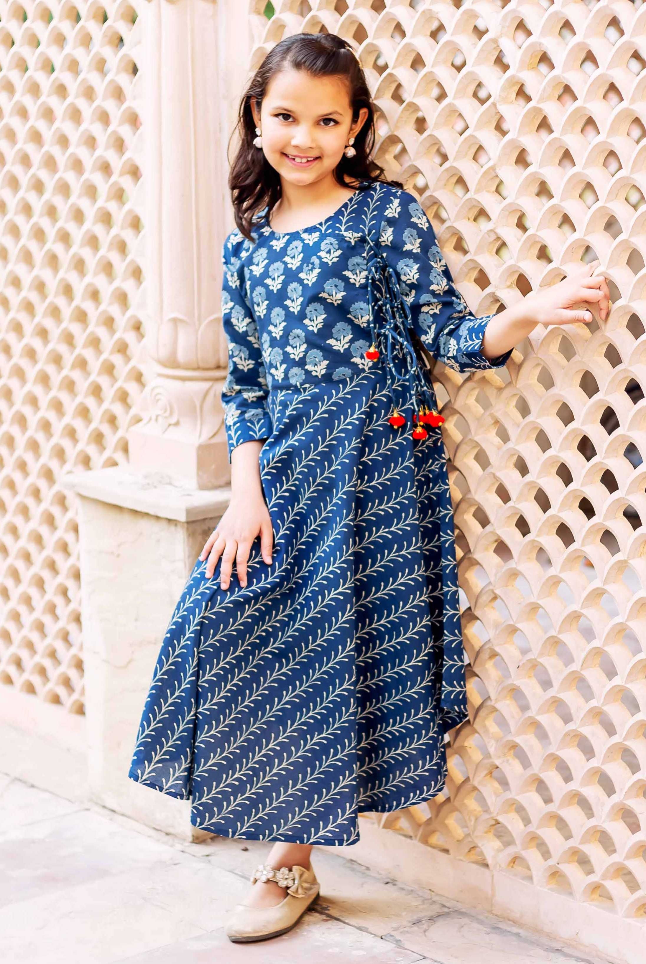 A young girl in a blue, floral-patterned dress with tassels, posing against a wall.