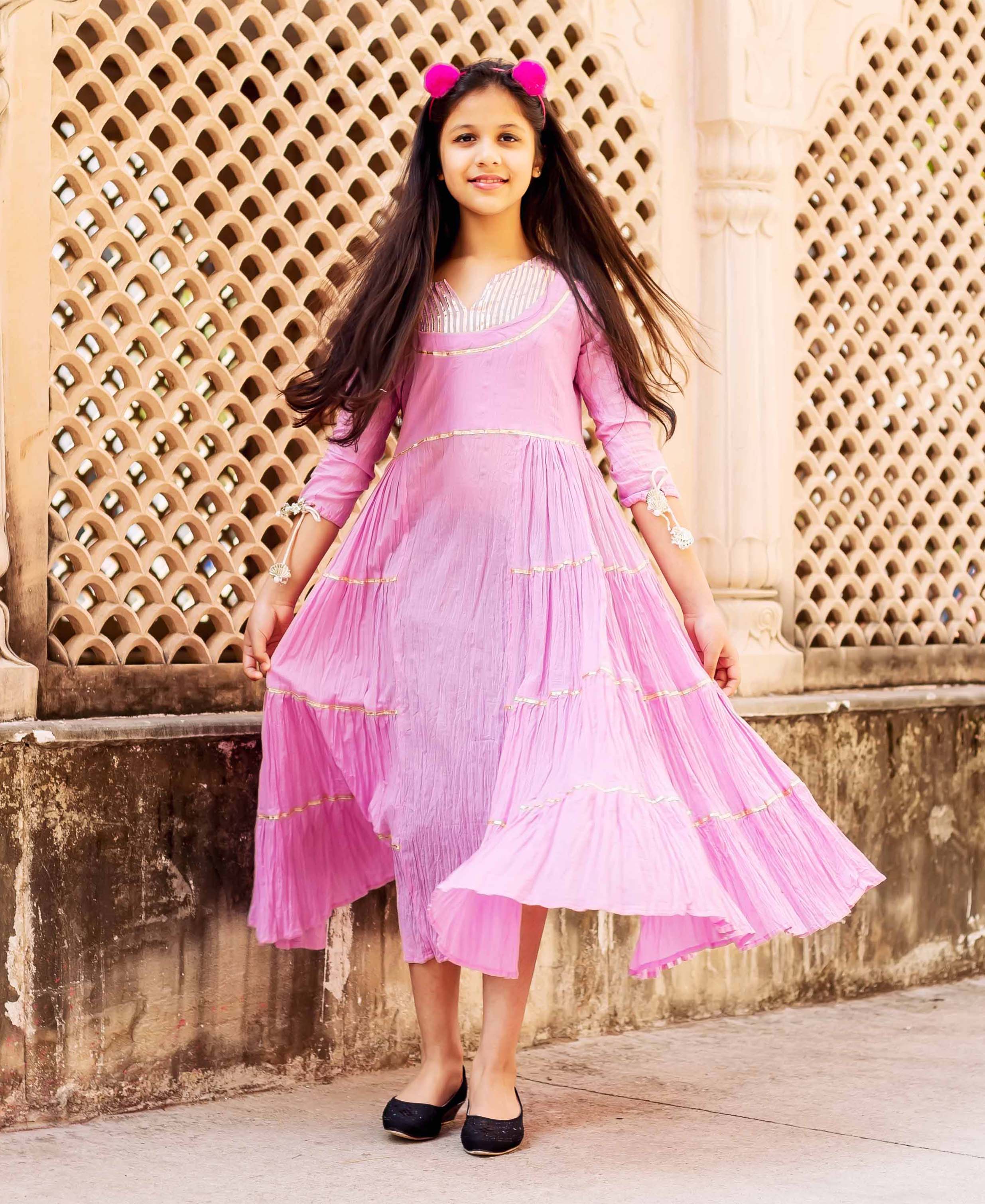 A young girl in a pink, long-sleeved dress with gold trim and a ruffled hem.