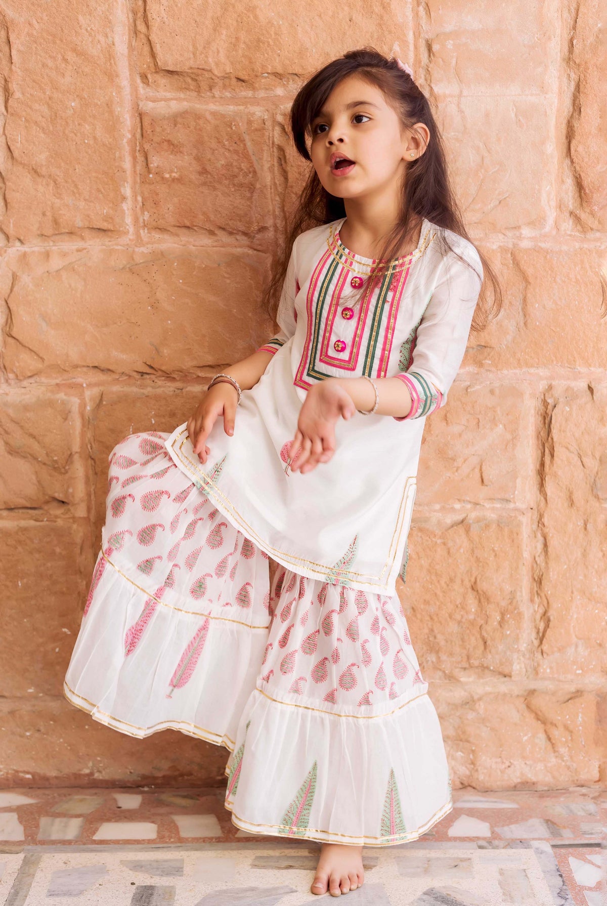 Young girl in white sharara set with pink, green, and gold block prints.
