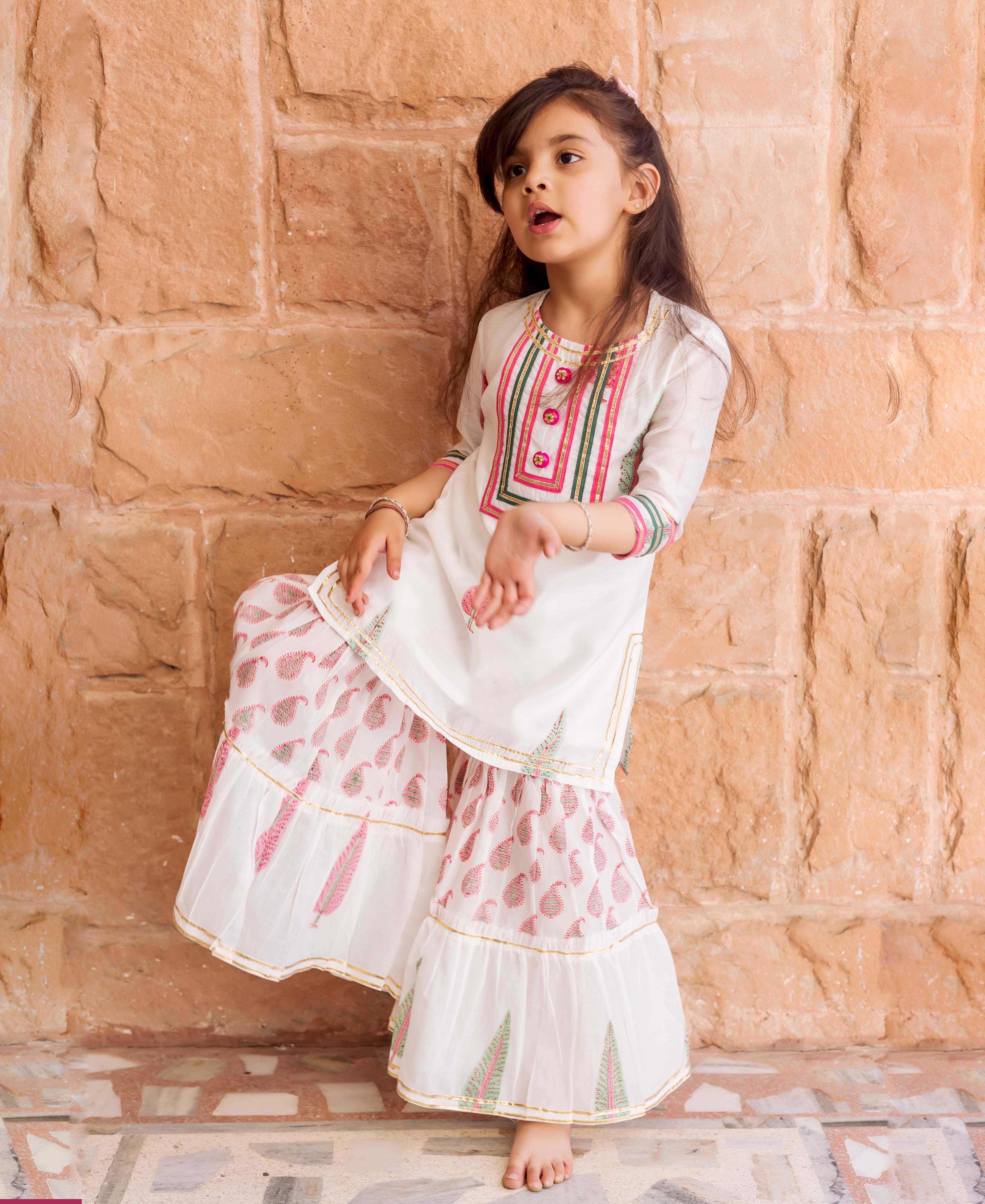 Young girl in white sharara set with pink, green, and gold block prints.