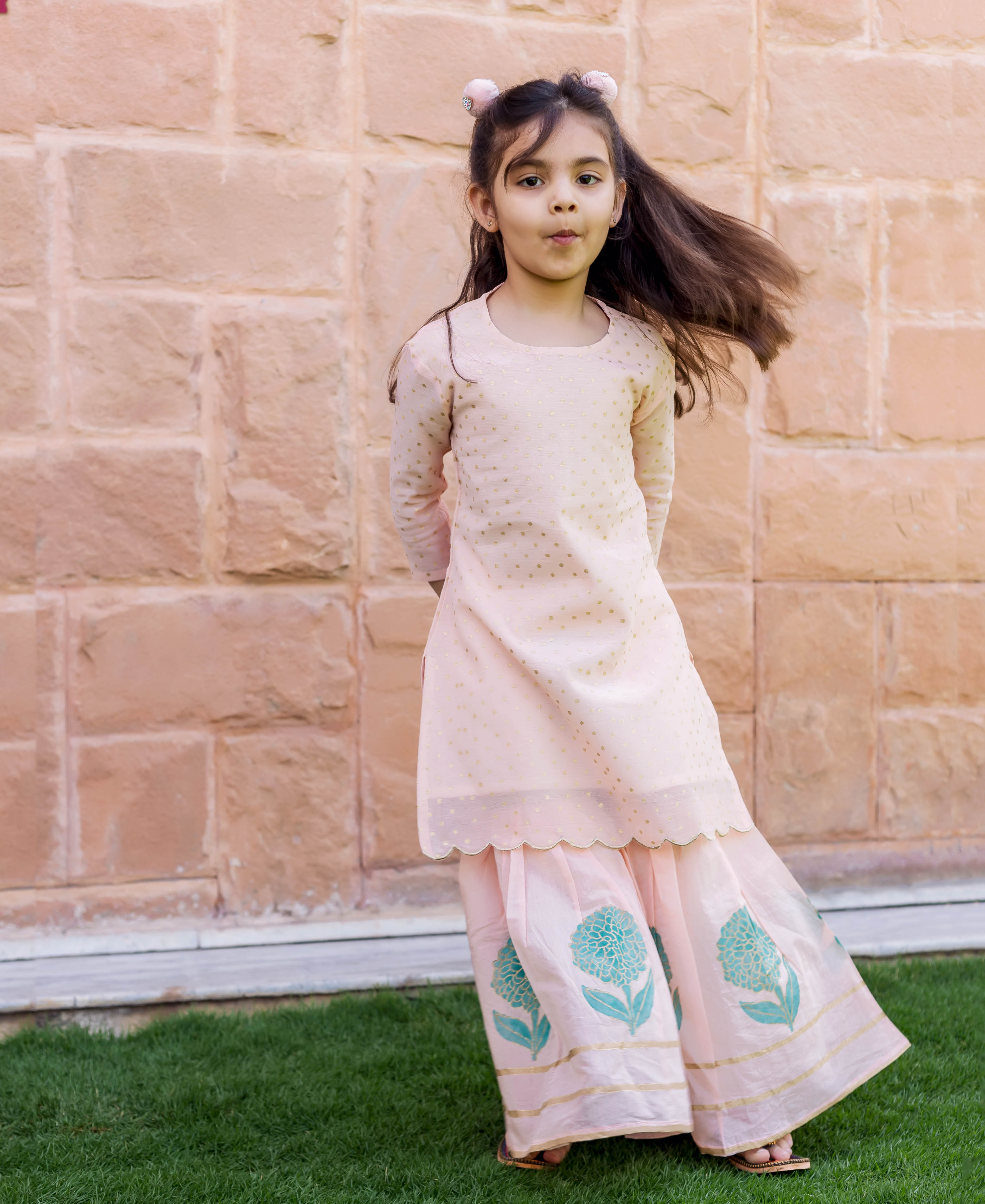 Full-length shot of a child in peach polka dot kurta and teal floral sharara set.