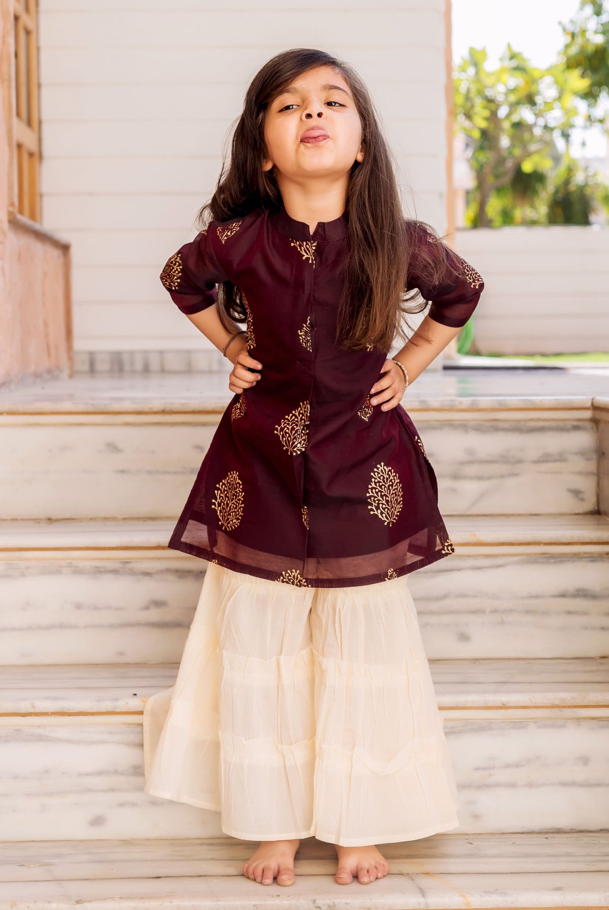 Young girl in wine kurta with gold print and crème tiered sharara standing on marble stairs.