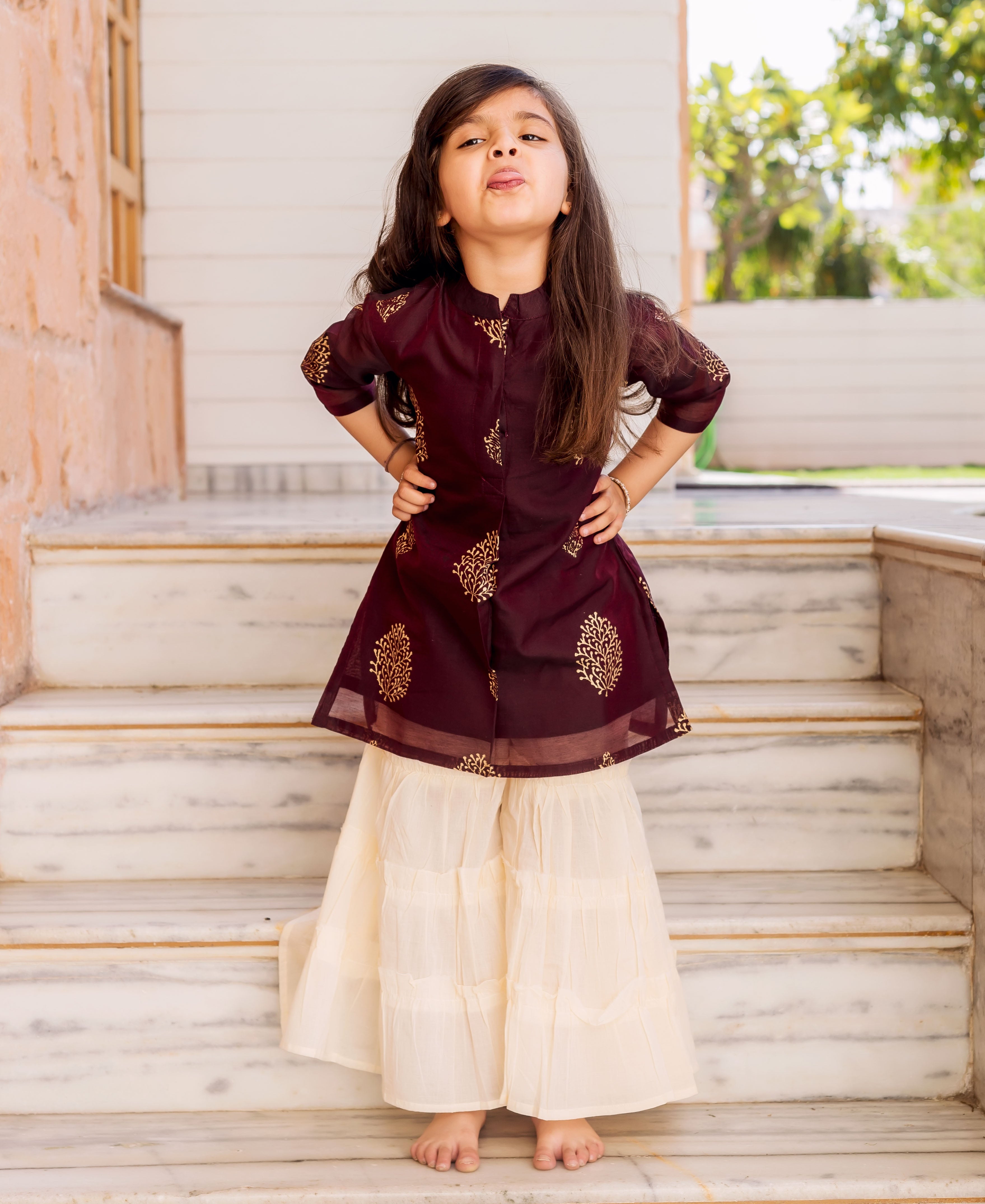 Young girl in wine kurta with gold print and crème tiered sharara standing on marble stairs.