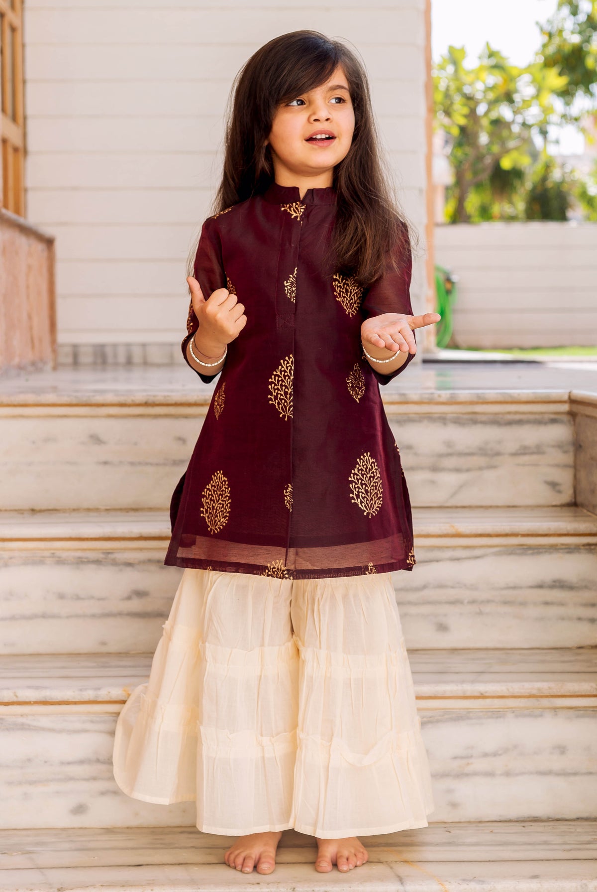Child model wears a wine gold-printed tunic with cream tiered sharara set.