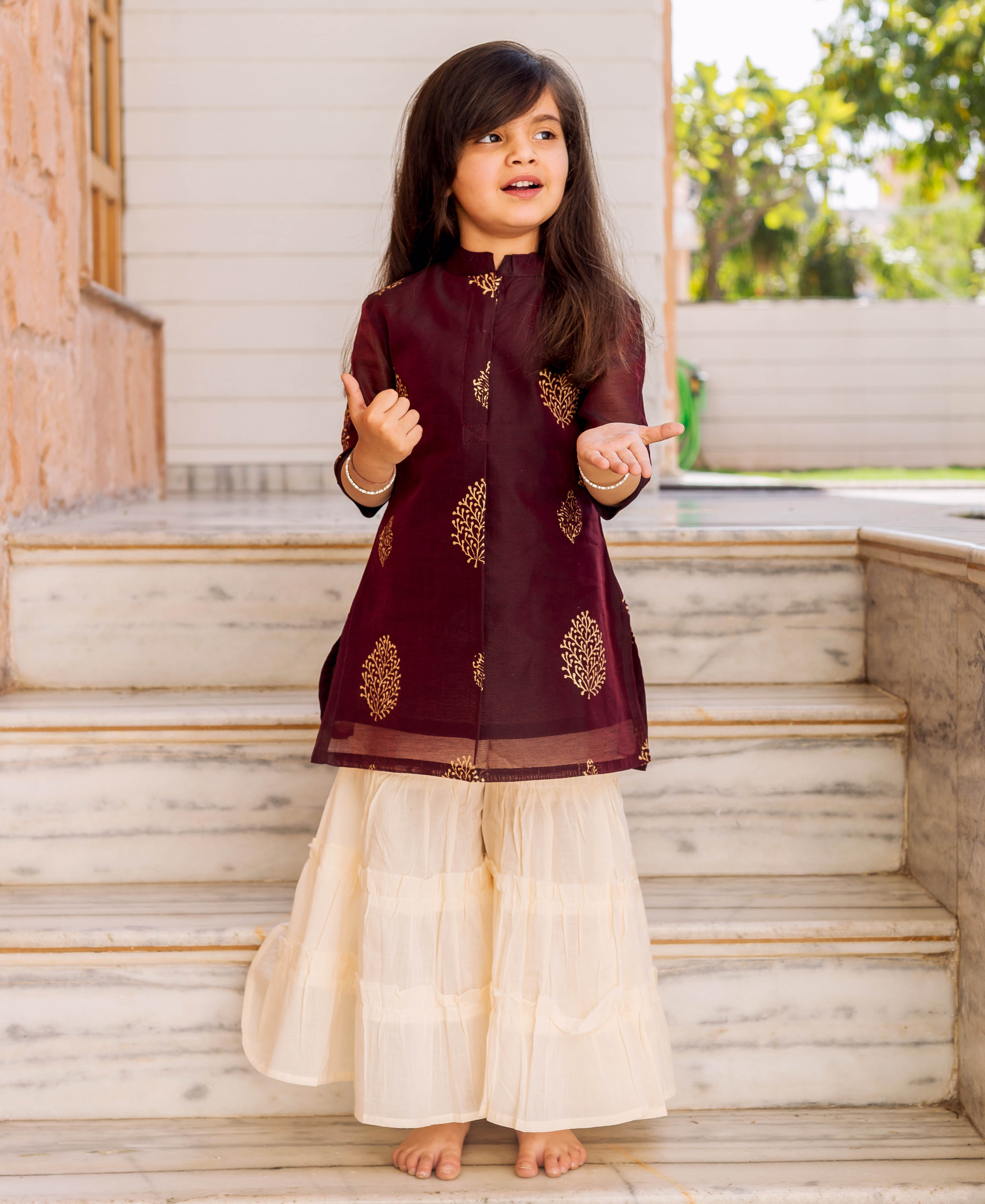 Child model wears a wine gold-printed tunic with cream tiered sharara set.