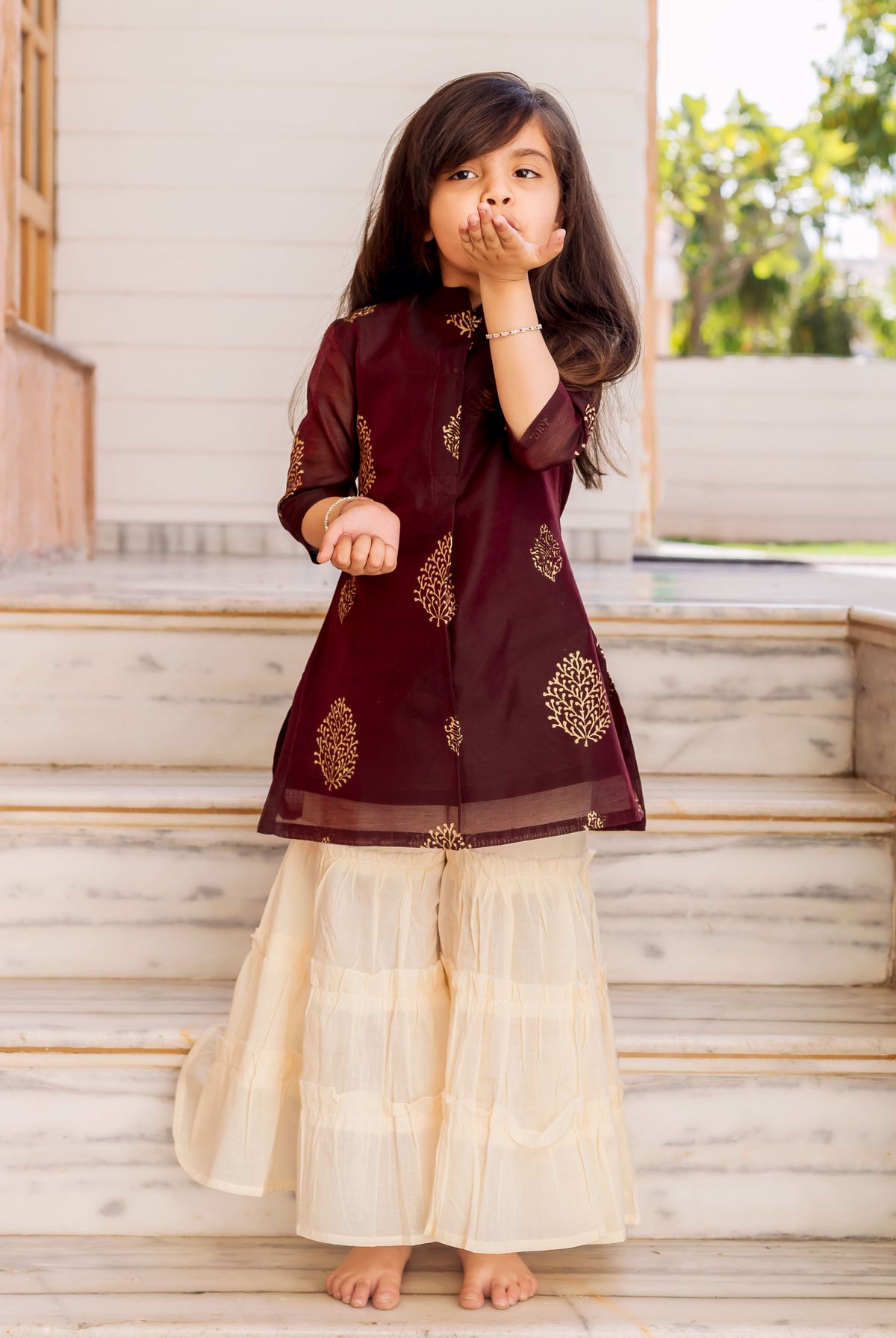 Little girl models a wine gold-printed kurta and crème sharara set on marble steps.