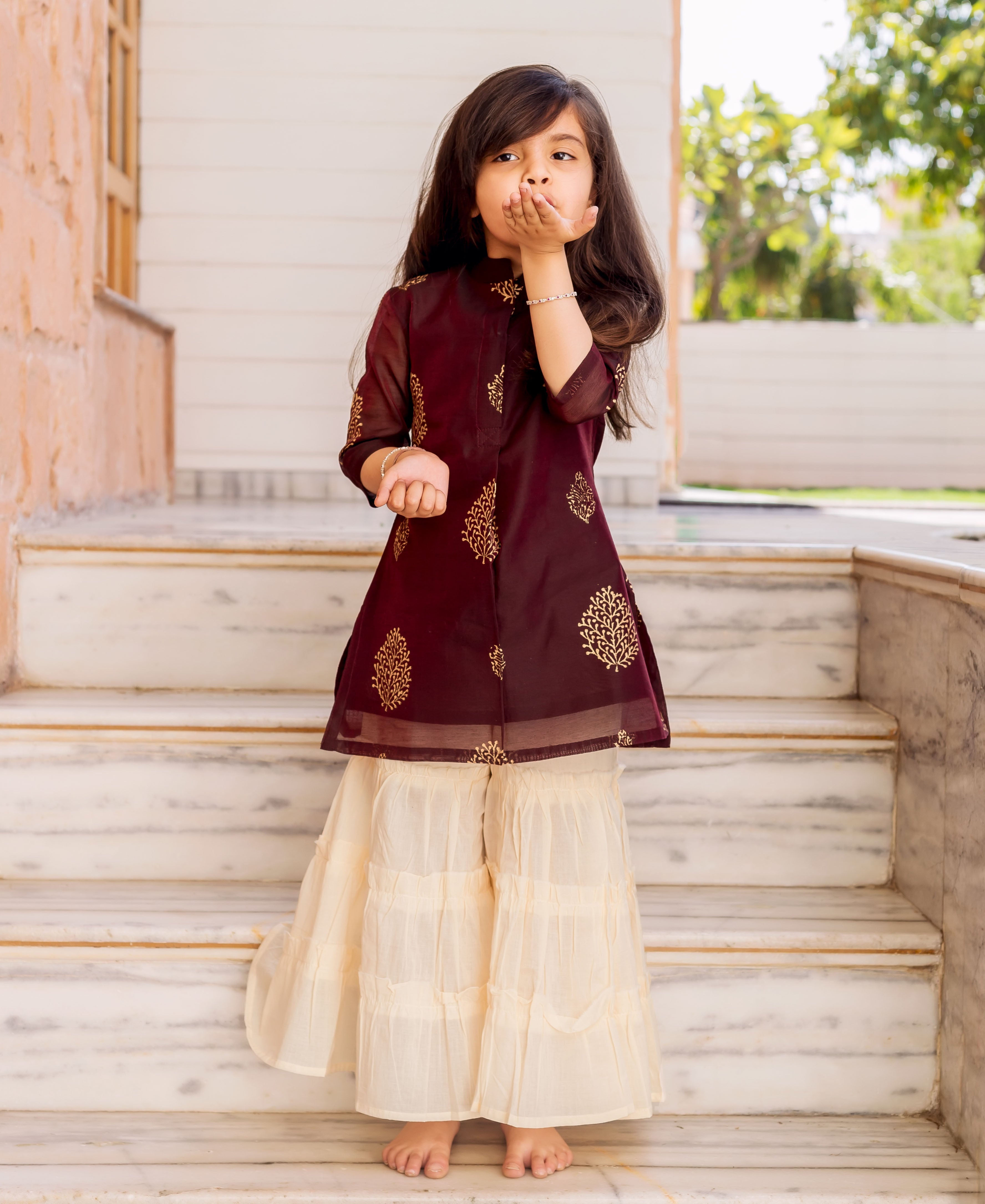Little girl models a wine gold-printed kurta and crème sharara set on marble steps.