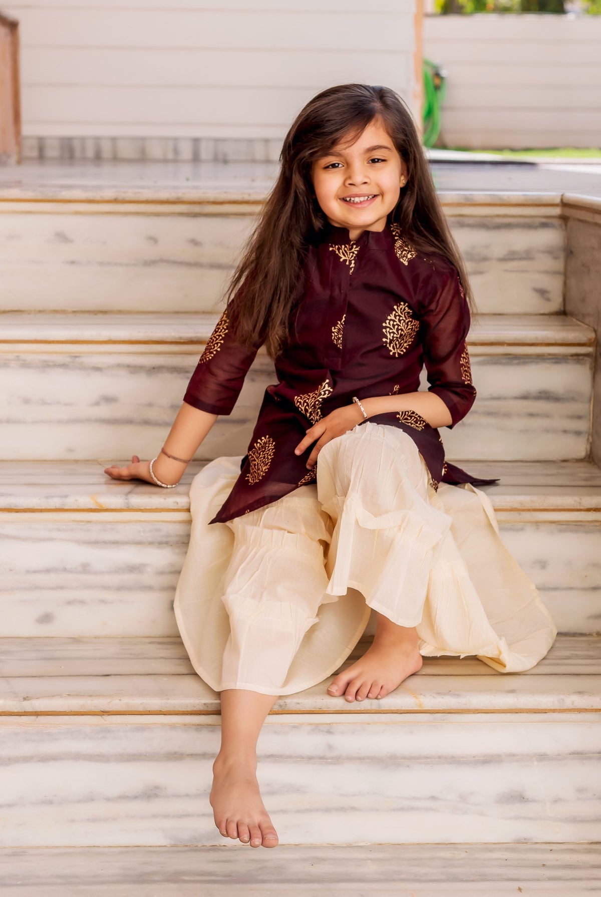Smiling girl wears a wine and crème gold printed sharara set, sitting on stairs.