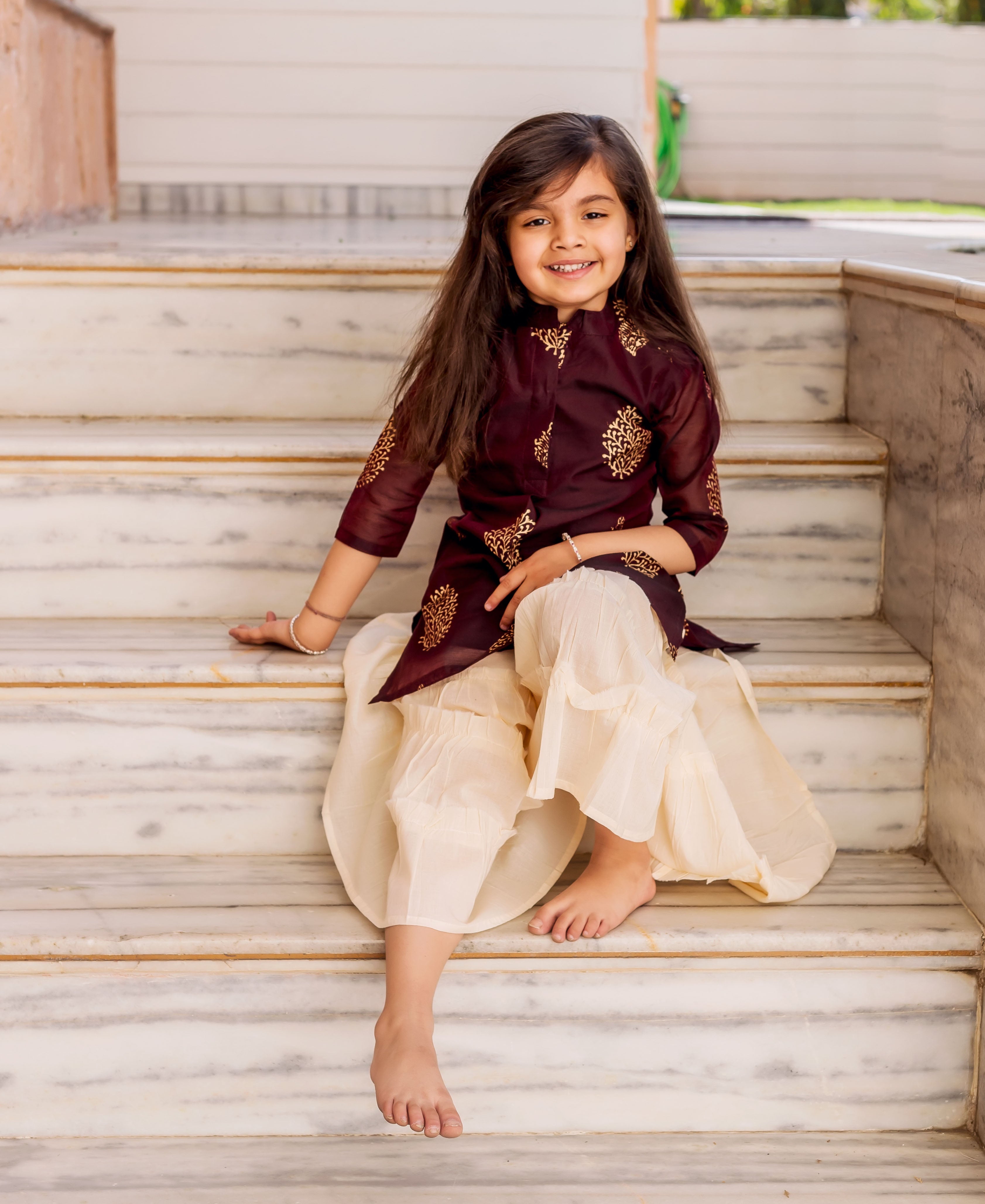 Smiling girl wears a wine and crème gold printed sharara set, sitting on stairs.