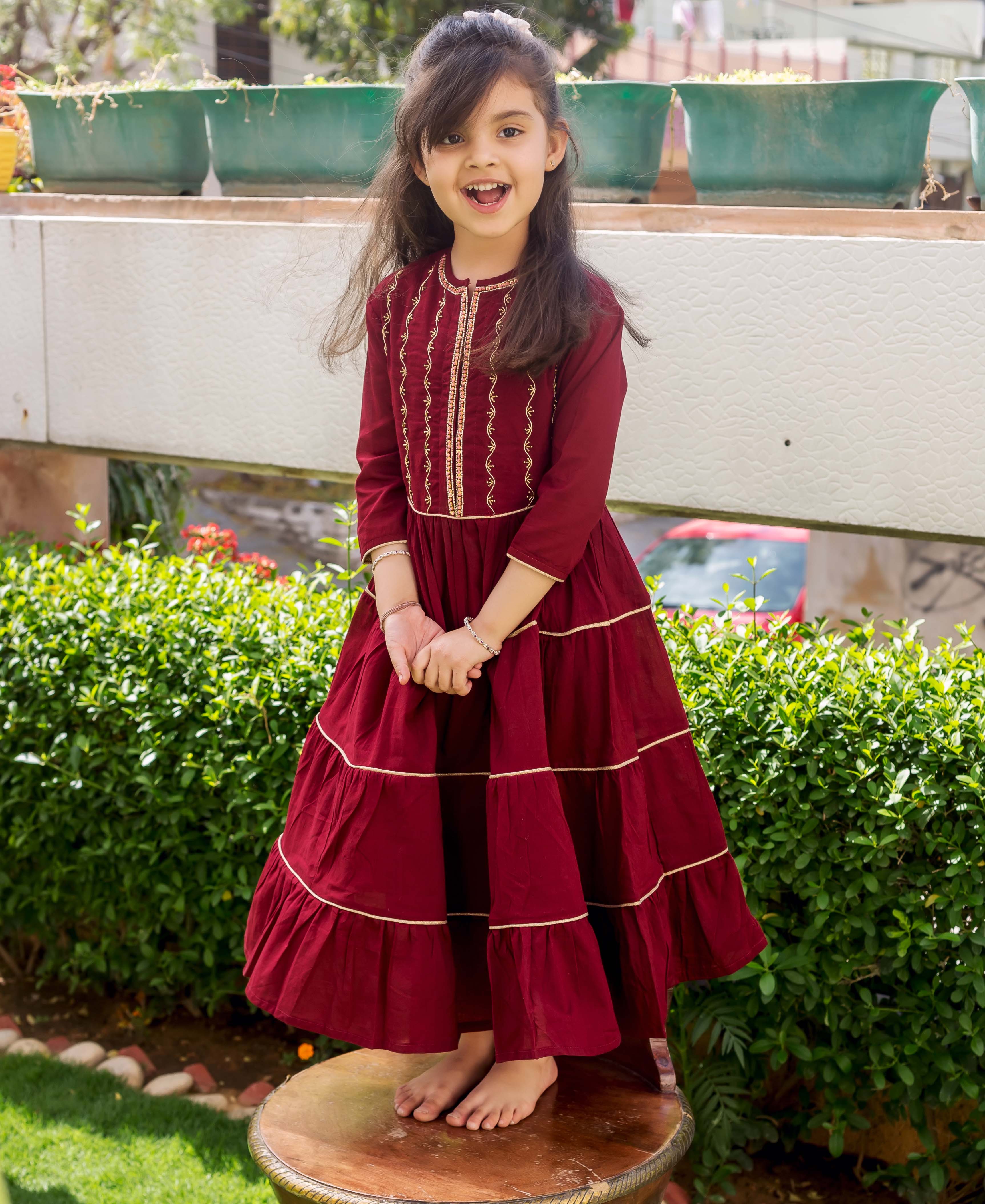 A young girl in a maroon, tiered, hand-embroidered dress with gold accents.