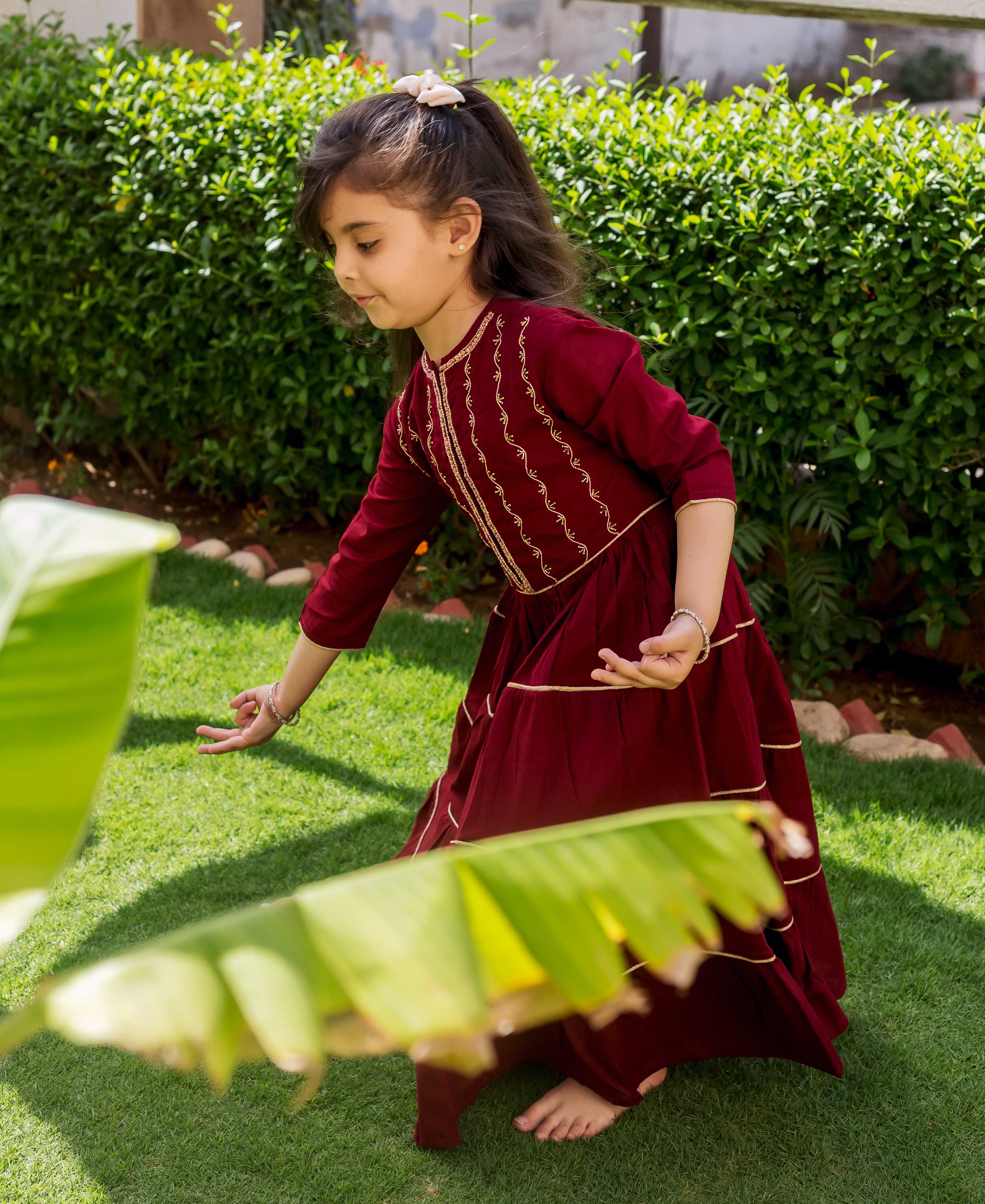 A young girl in a maroon dress with gold embroidery, standing on grass.