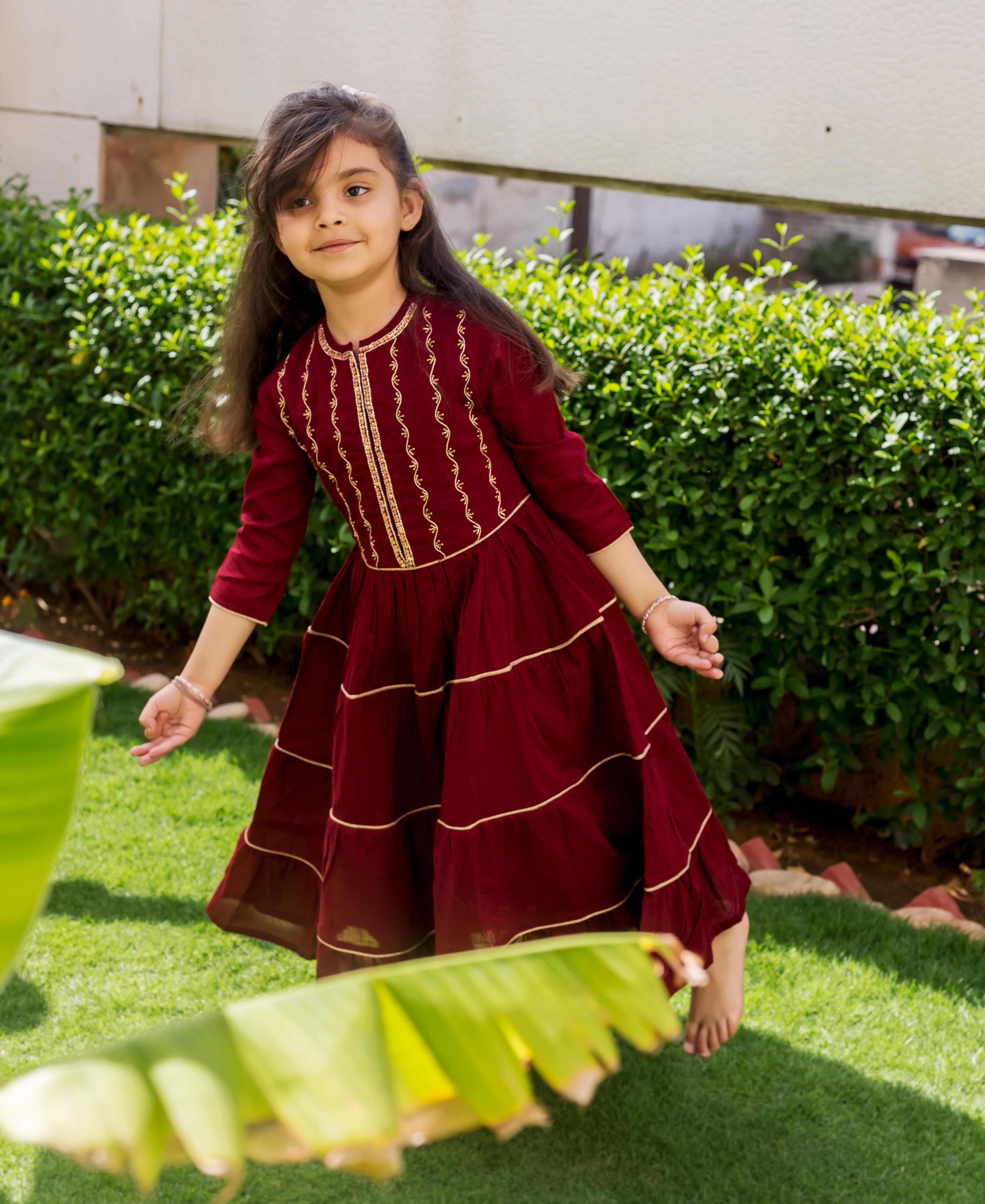 Maroon tiered dress with golden hand embroidery and trims, on a young girl in a garden.