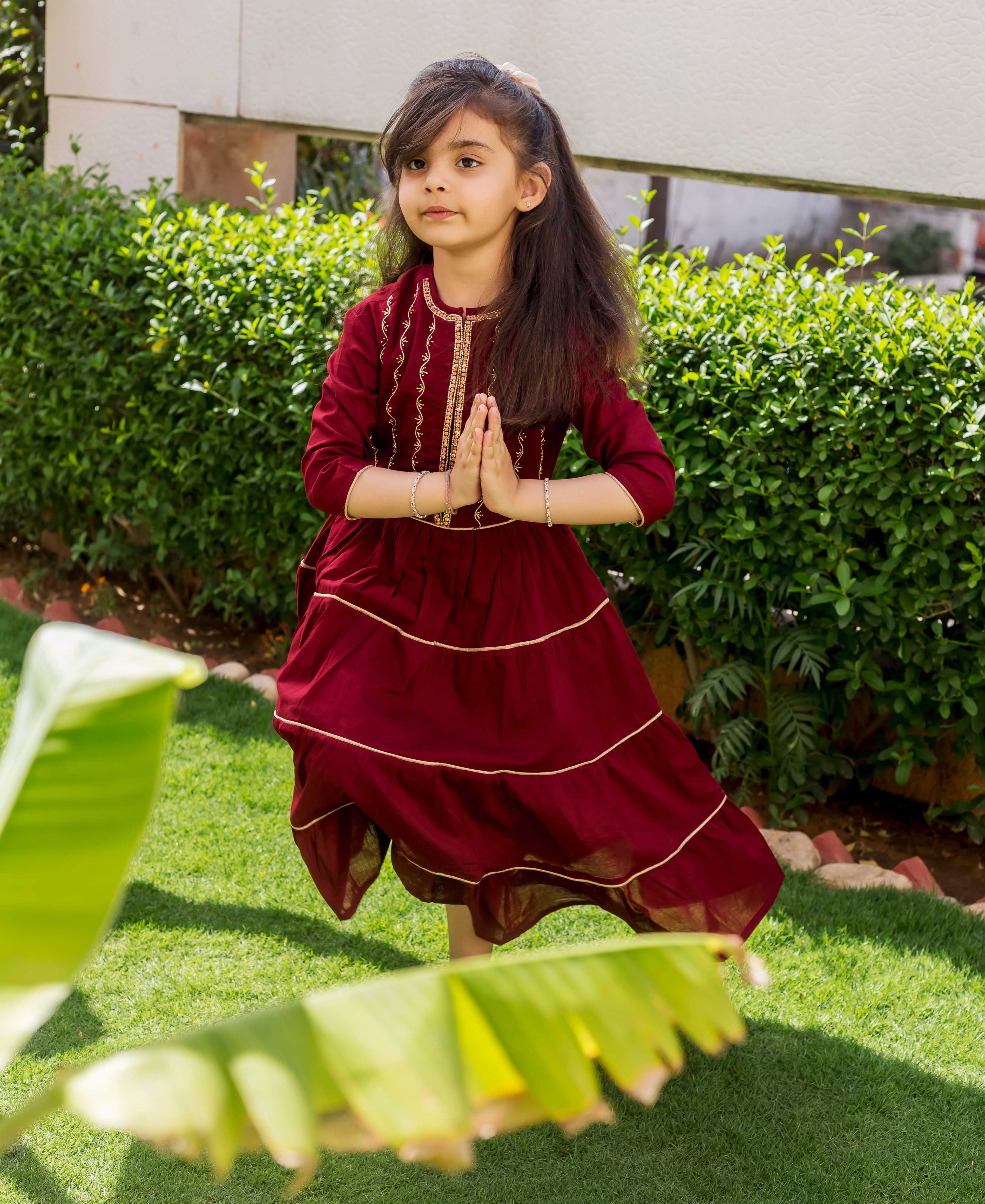 Young girl in maroon tiered dress with gold hand embroidery, standing in a garden.