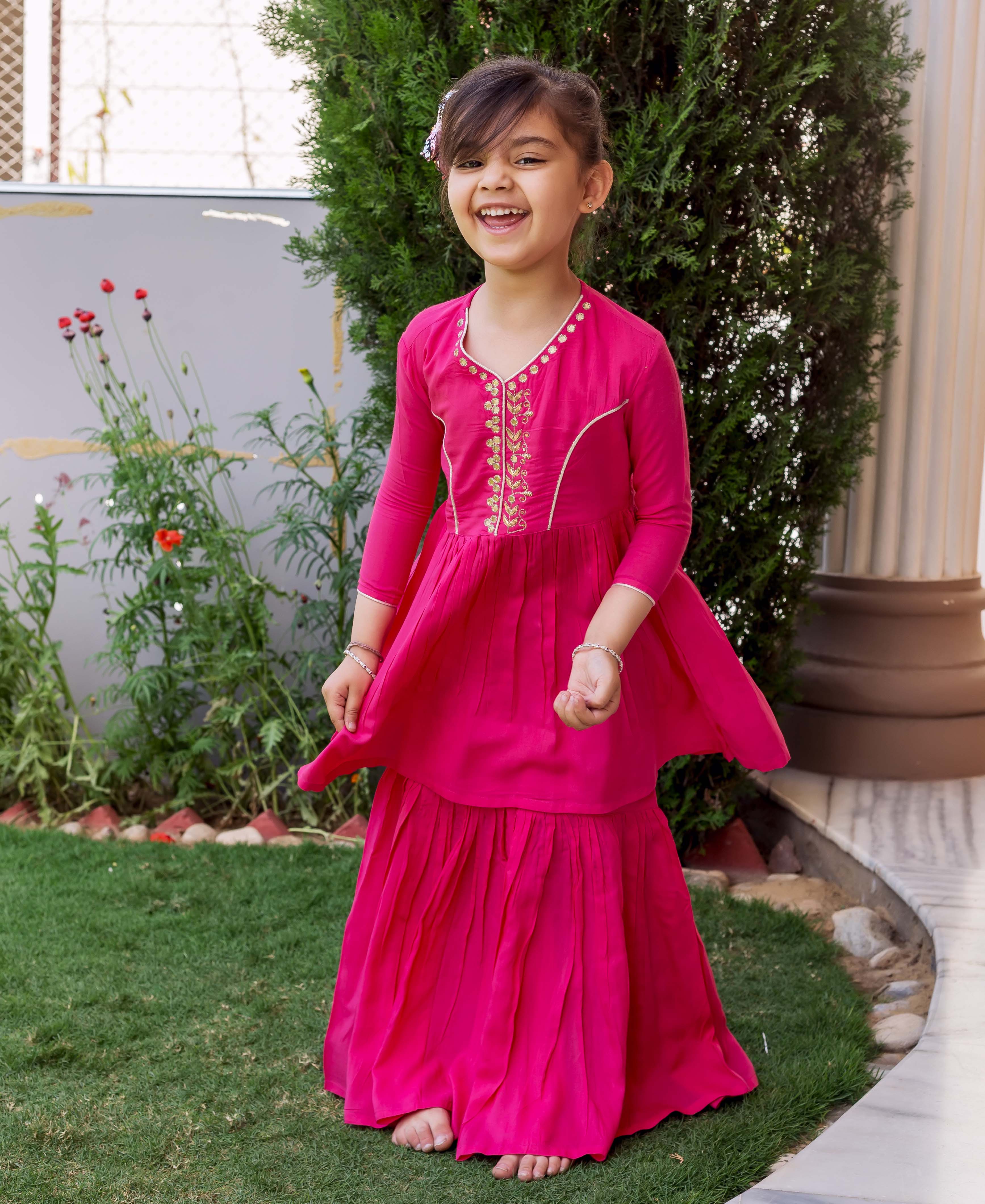 Vibrant pink hand-embroidered sharara set with gold details on a young girl outdoors.