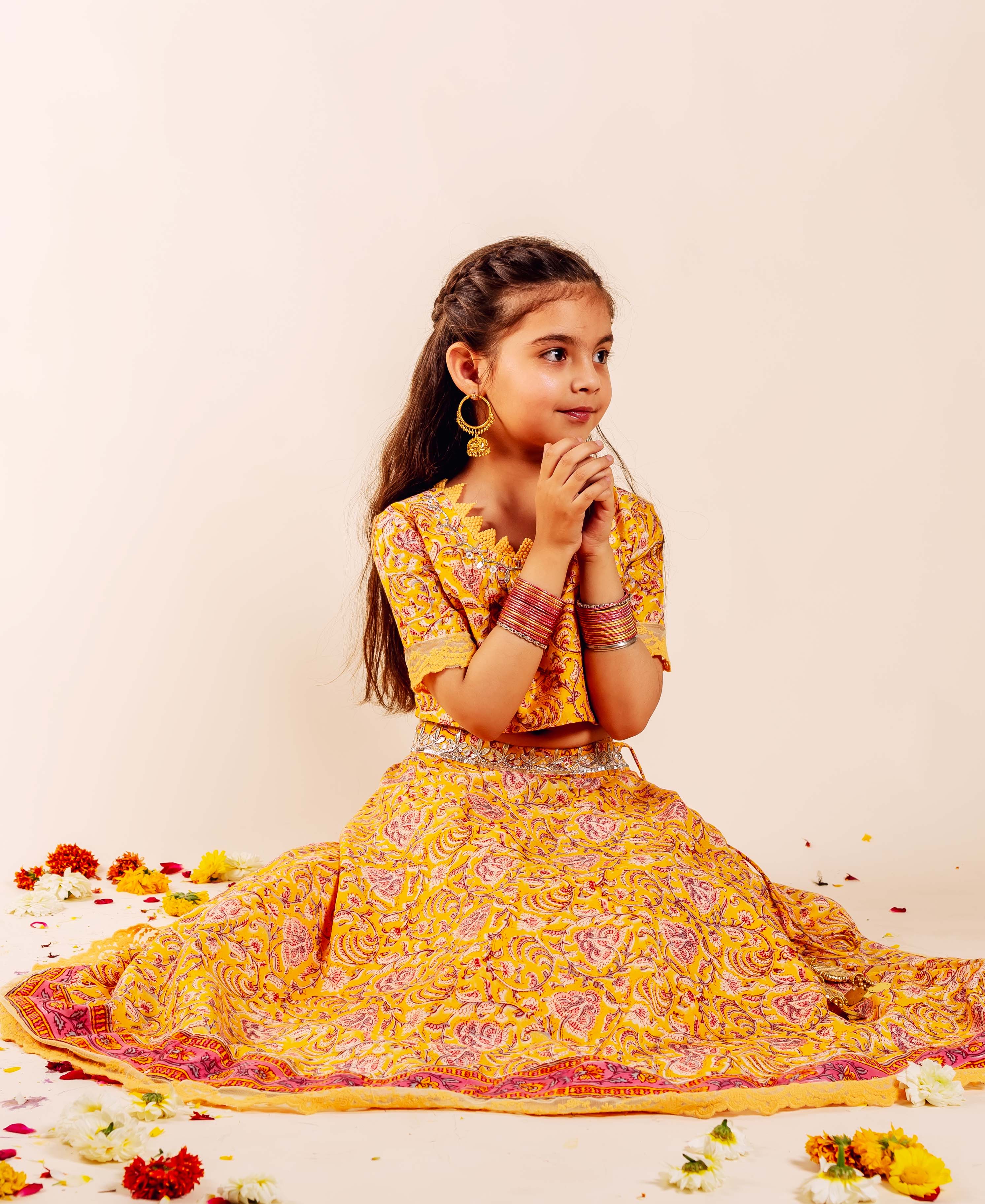 Baby girl in yellow hand-block-printed lehenga set, sitting with hands clasped.