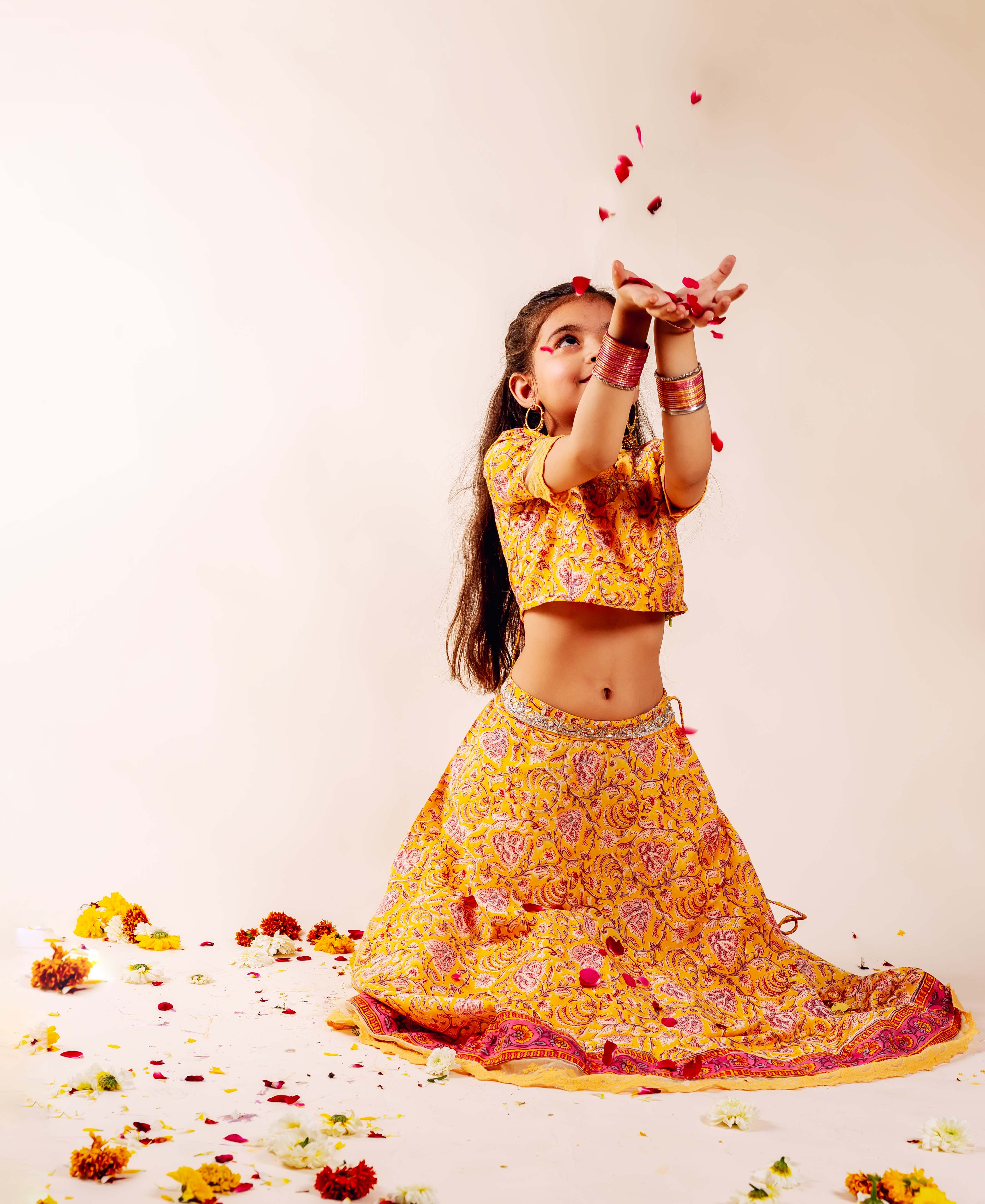 Child model in yellow hand-block print Farida Lehenga Set with pink motifs, scattering rose petals.