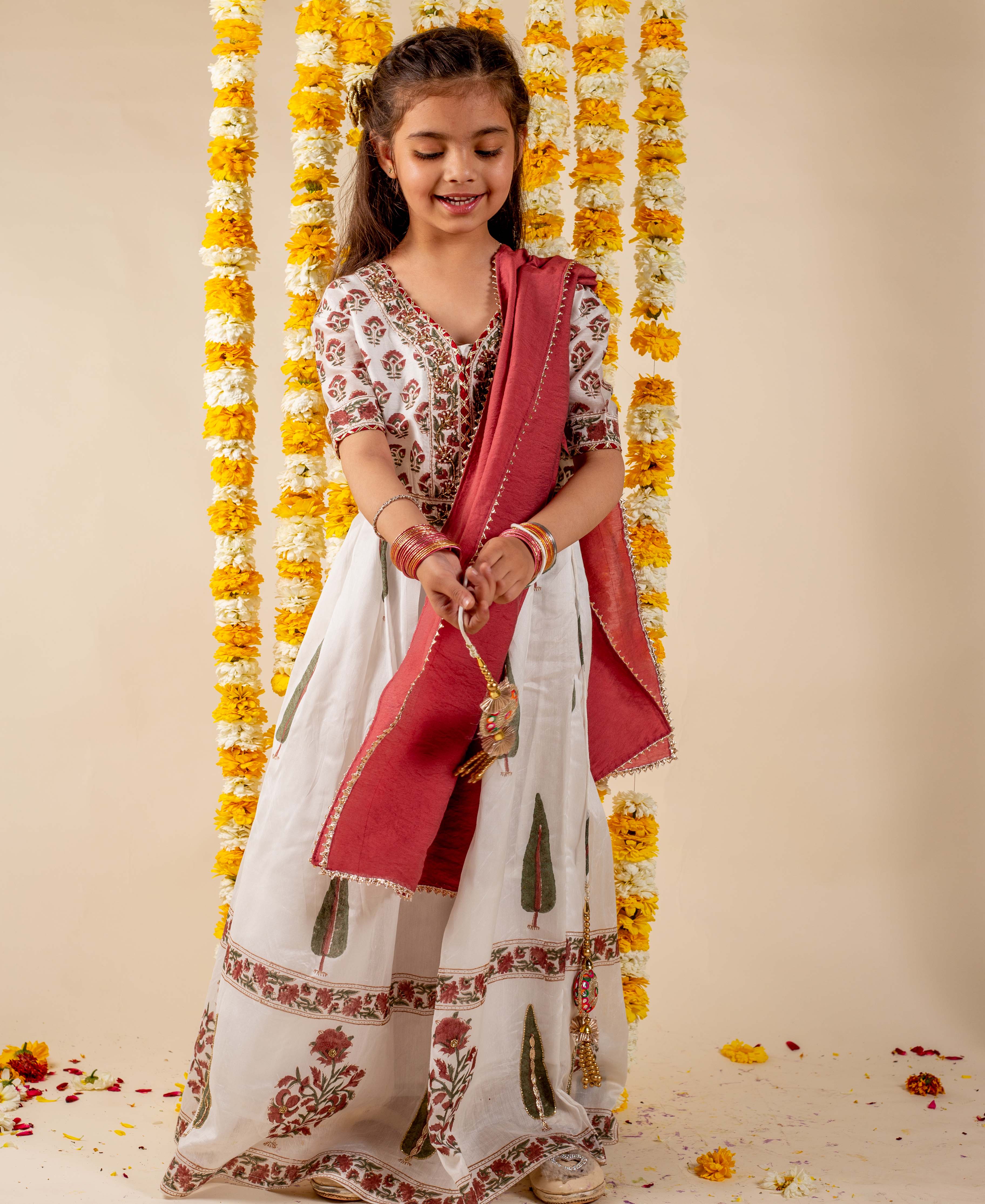 Smiling girl in white hand-block-printed lehenga set with red dupatta, green tree and floral patterns.