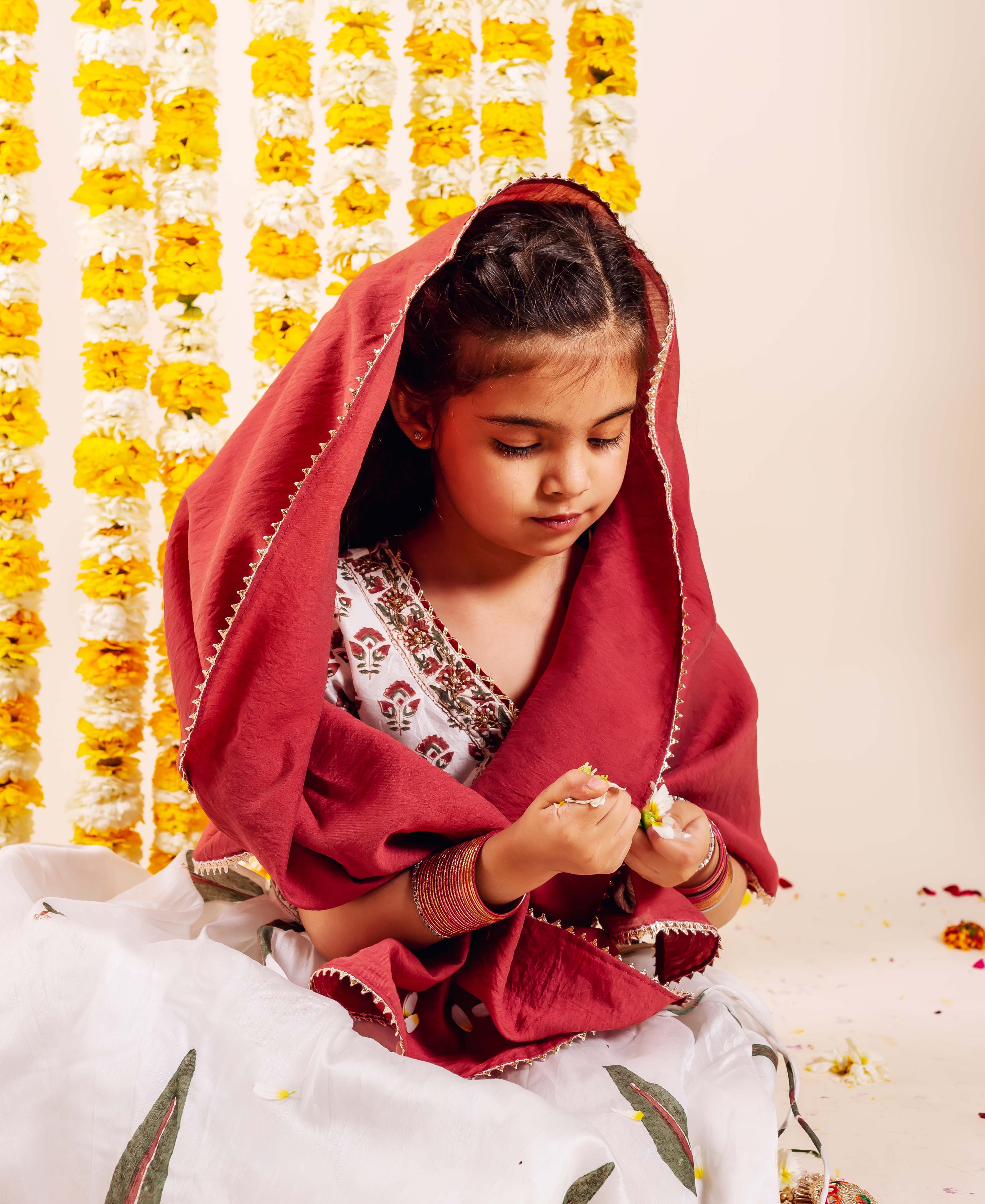 Baby girl wearing a white Nayab lehenga set with green leaf print, red dupatta.