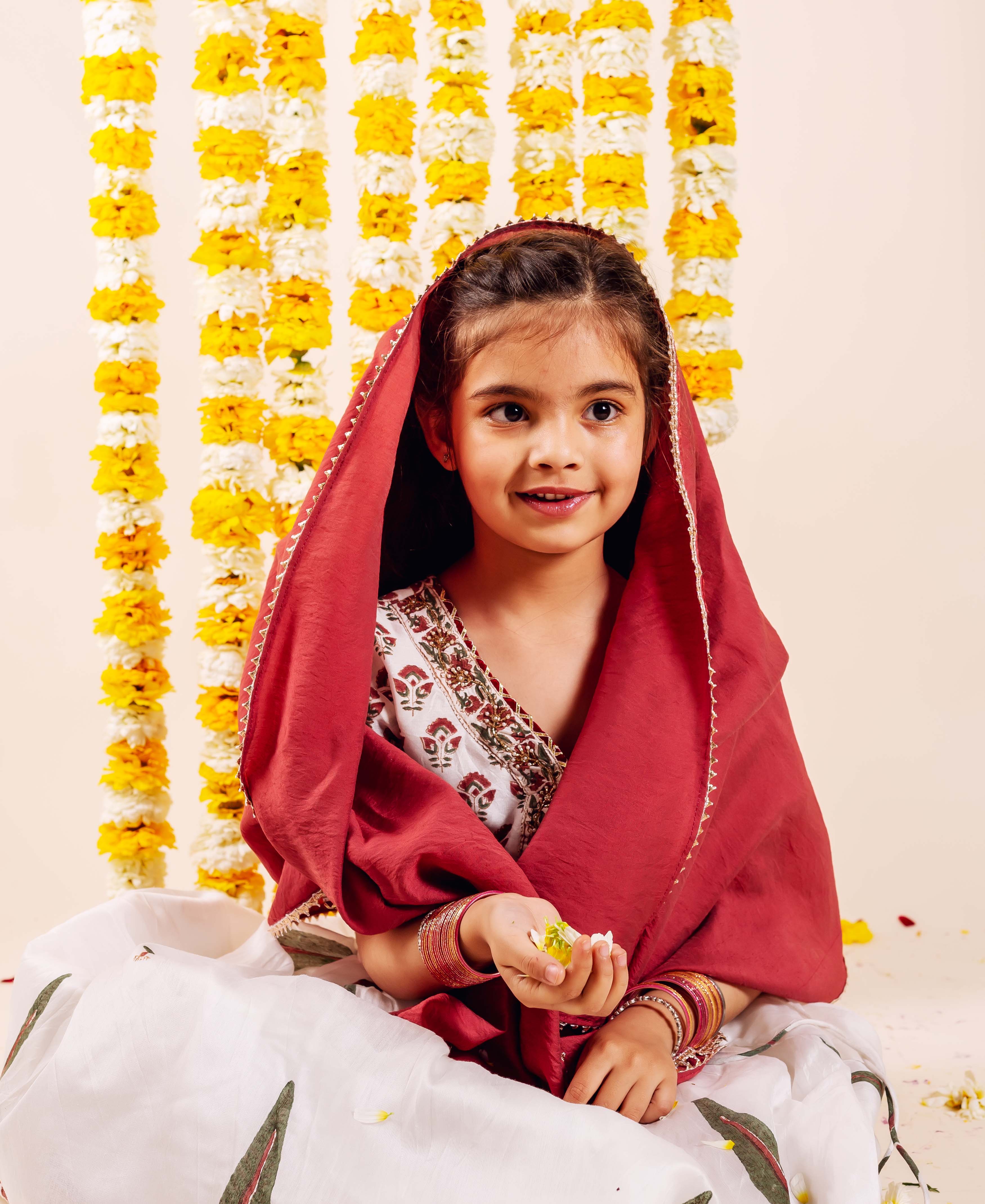 Smiling girl in a red dupatta and white hand-block-printed lehenga set, holding flowers.