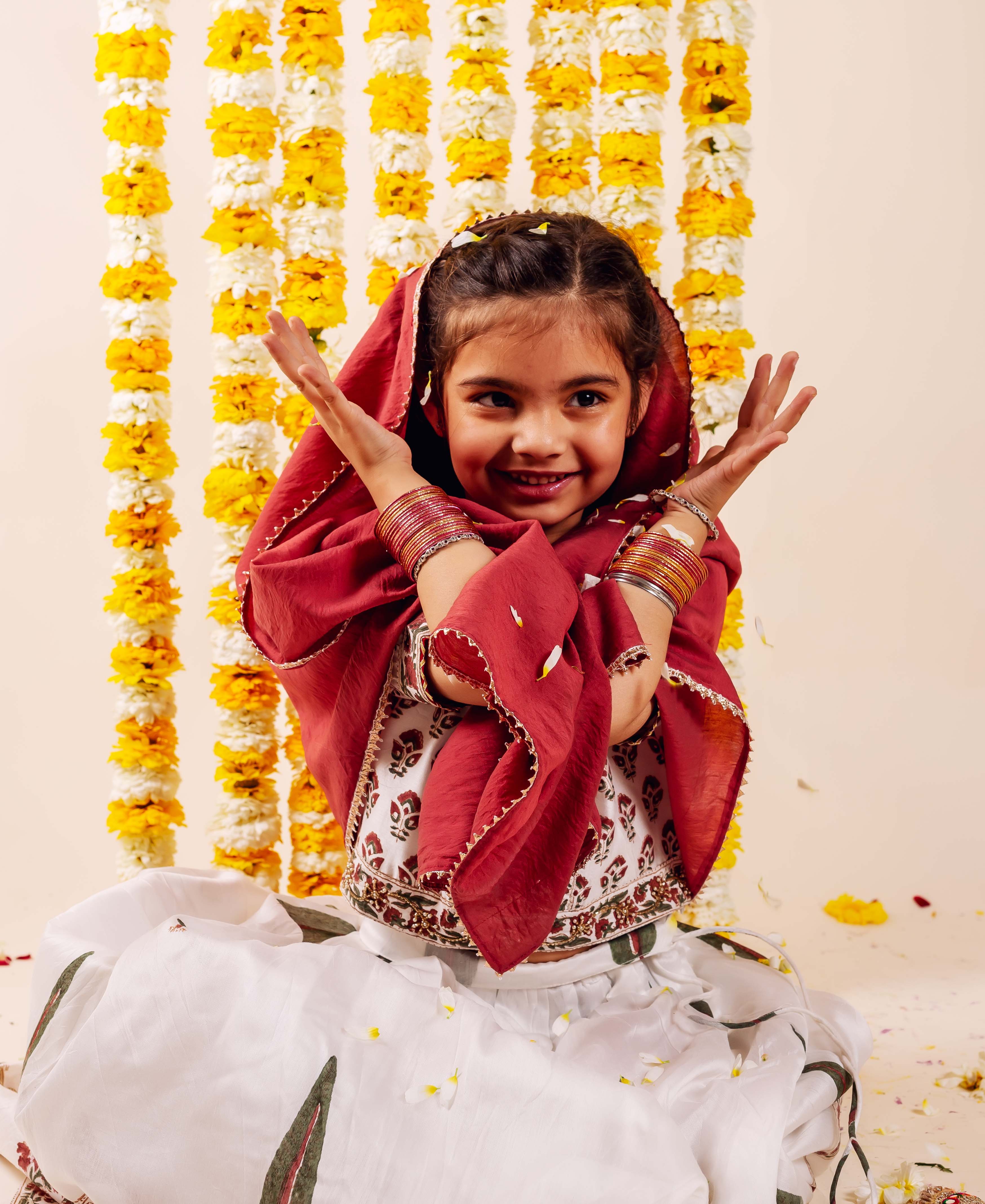 Smiling baby girl wears red dupatta, white embroidered hand-block printed lehenga set with bangles.