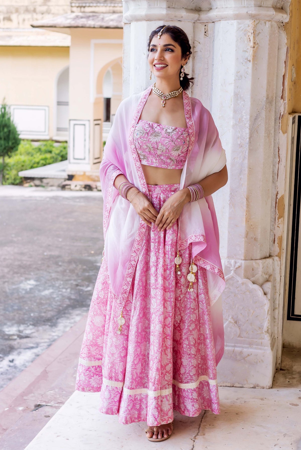 Woman in pink floral lehenga set with white embroidery and sheer shawl standing outdoors.