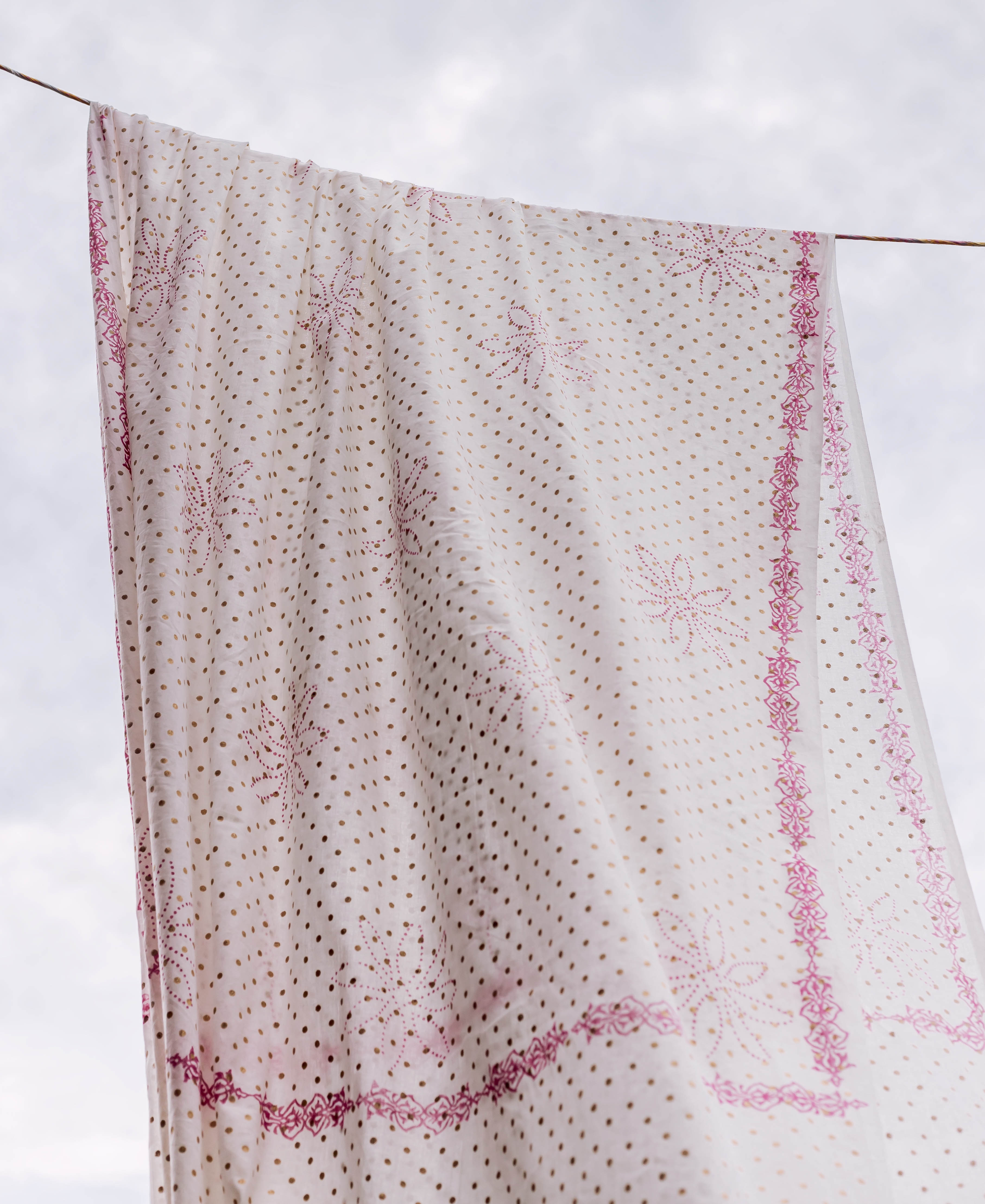 White dupatta with pink block-printed patterns and gold polka dots, softly draped outdoors.