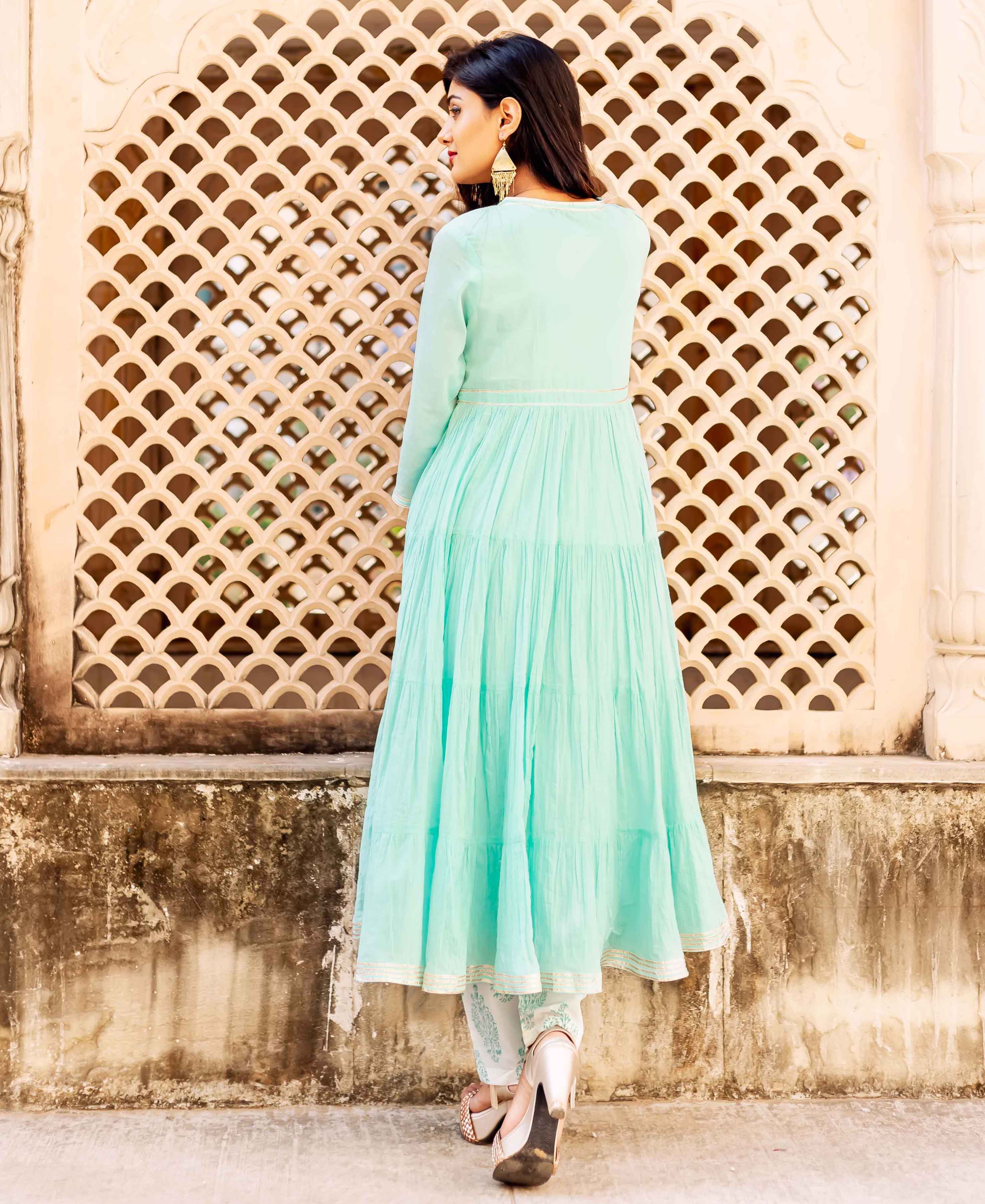 Woman in seafoam green long dress with gold accents, standing in front of lattice wall.
