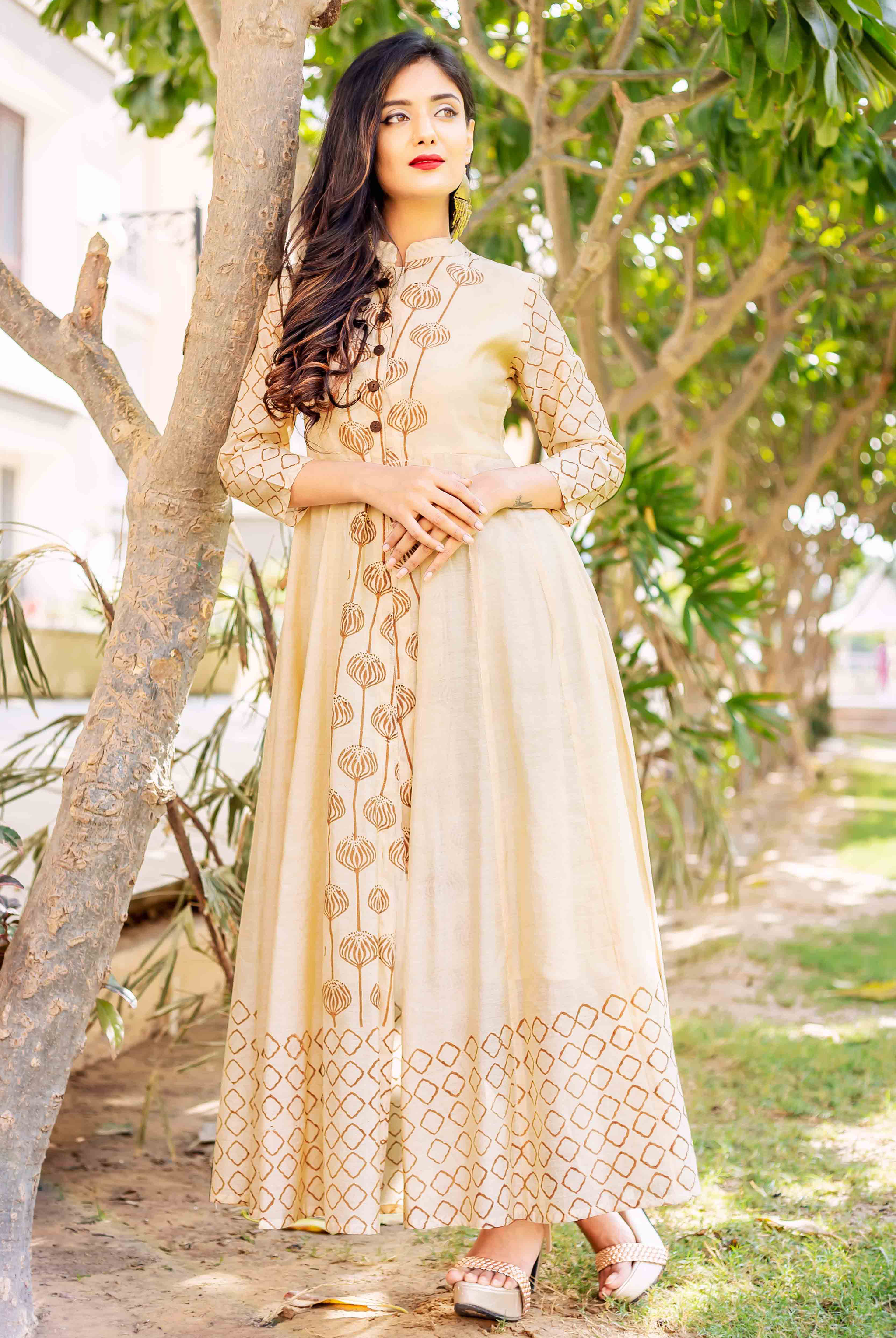 A model wearing a beige and brown printed dress with a tree in the background.