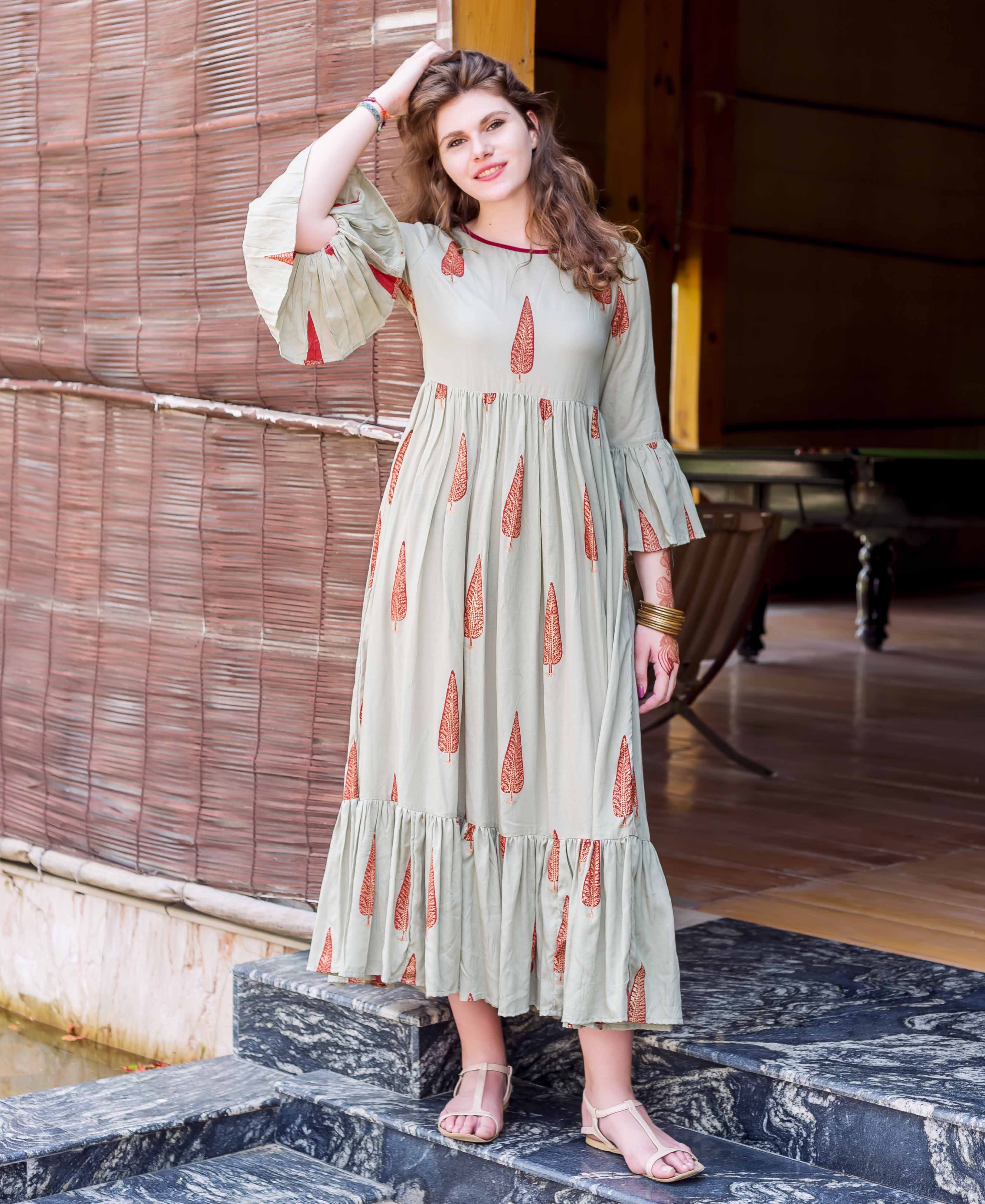 Model wears a sage green maxi dress with red block-printed leaf patterns, bell sleeves, and ruffled hem.