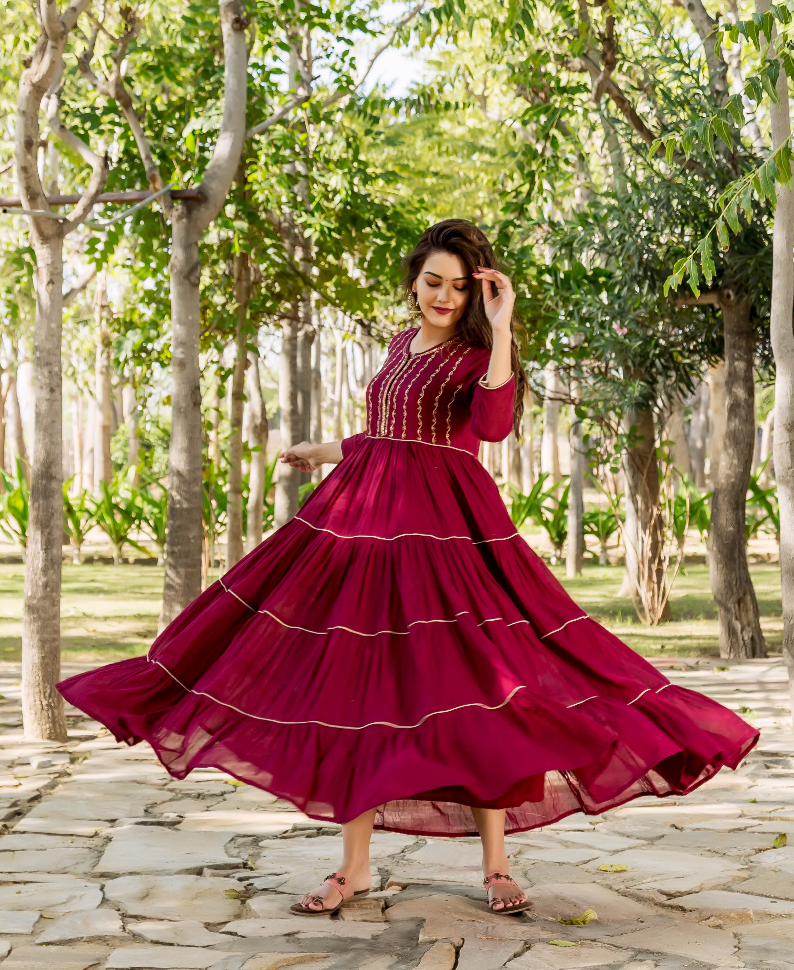 A woman in a maroon tiered dress with gold embroidery, posing outdoors.
