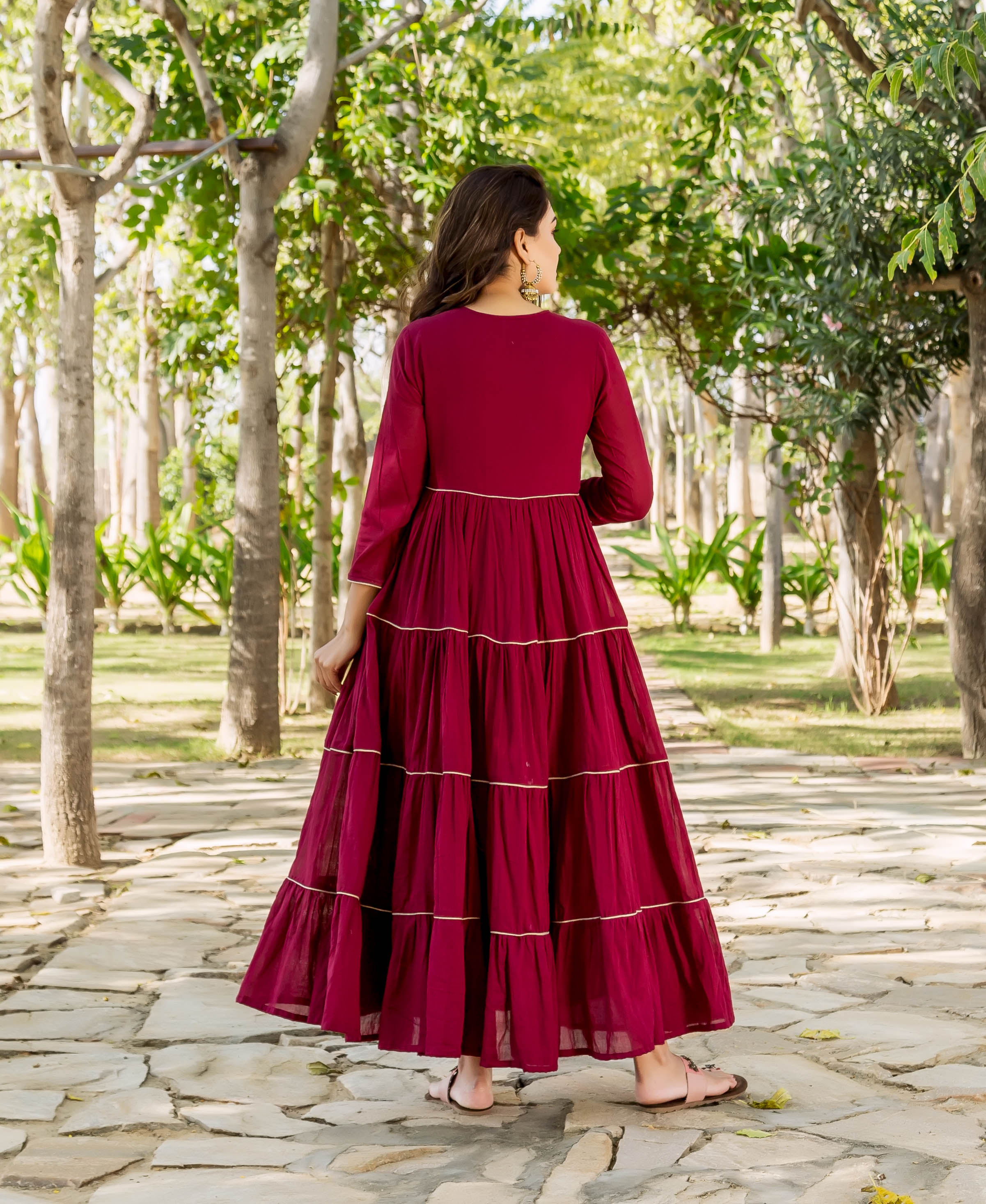 Maroon tiered dress on model, back view, featuring golden embroidered trim outdoors.