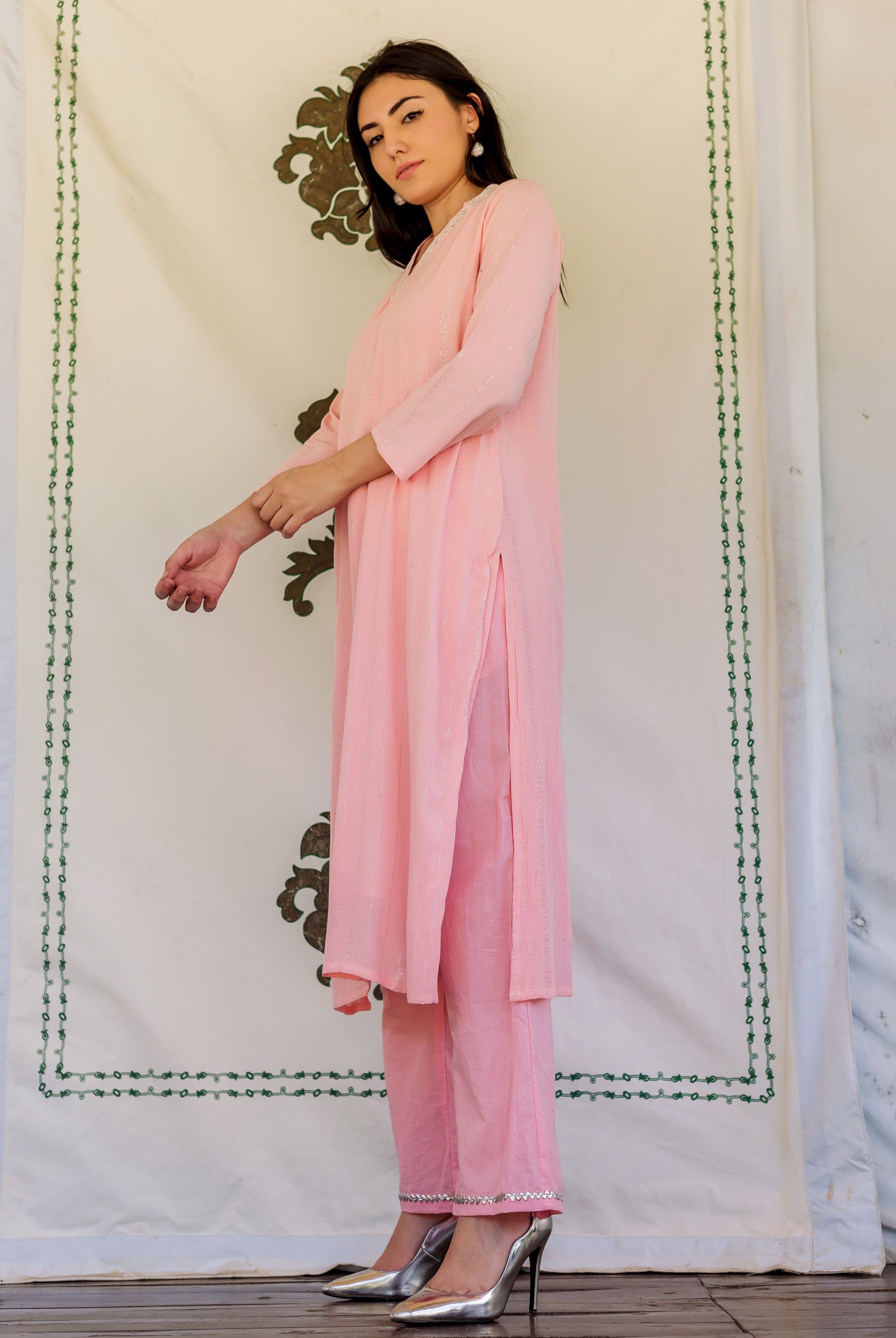 Woman wearing pink embroidered kurta and pants, posing in front of white fabric backdrop.