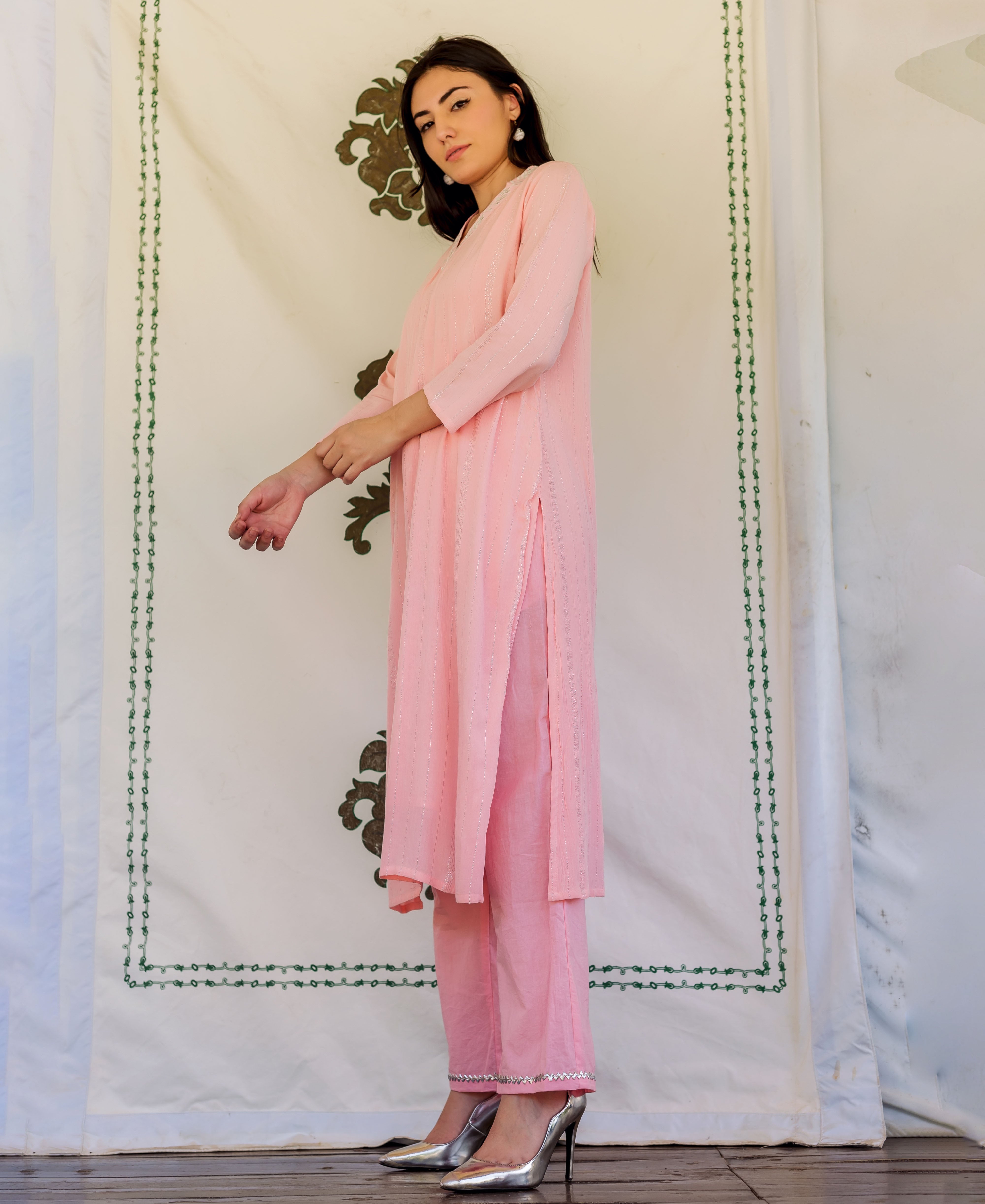 Woman wearing pink embroidered kurta and pants, posing in front of white fabric backdrop.