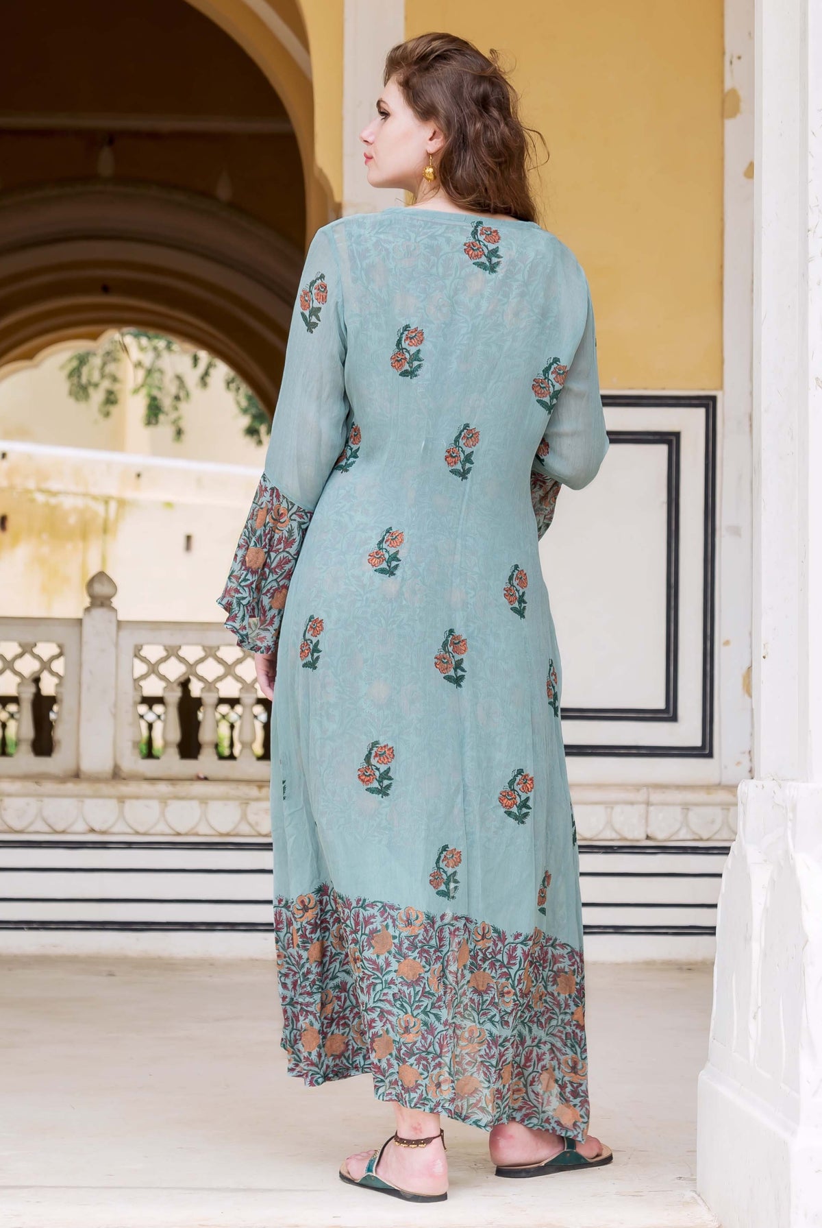 A model wearing a long, light blue dress with floral patterns and a matching jacket.