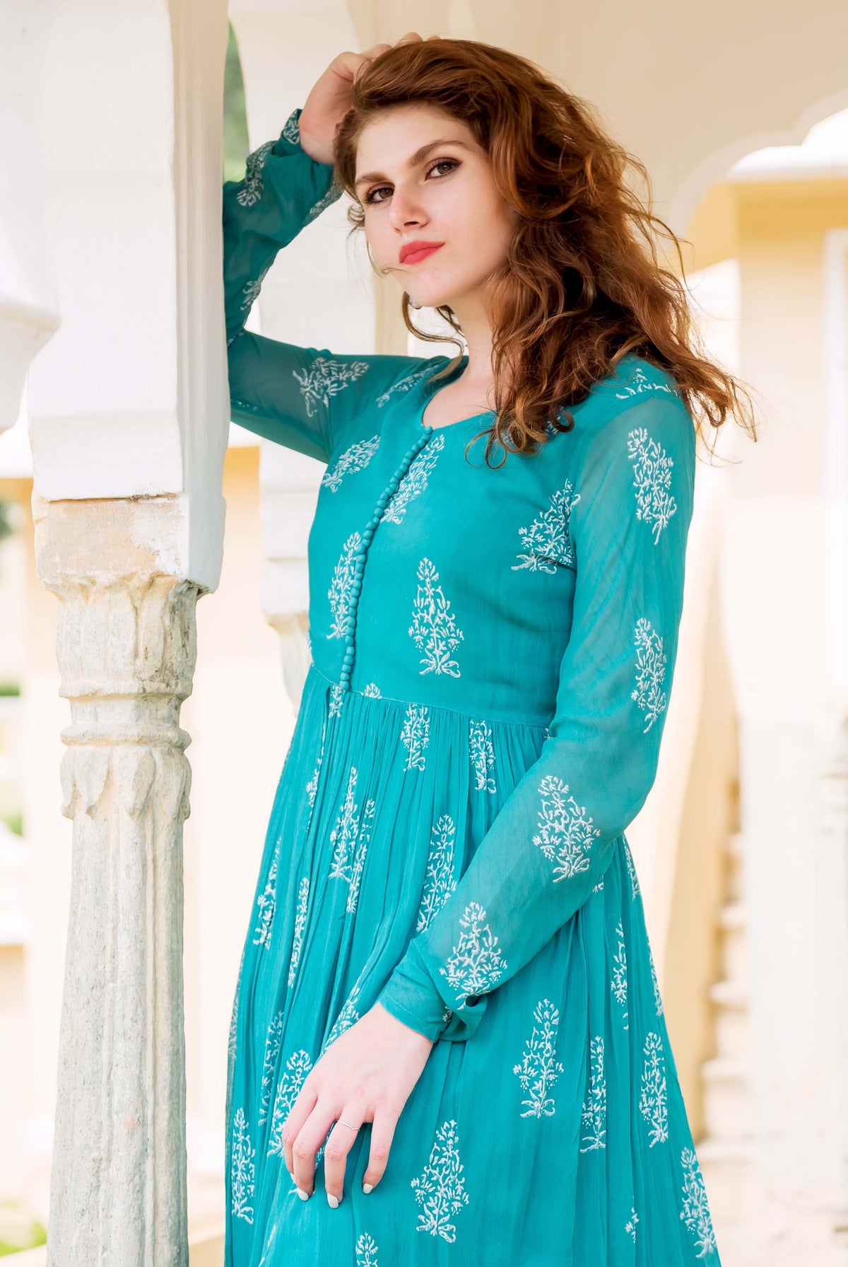 A woman in a teal dress with white block prints, posing beside a column.