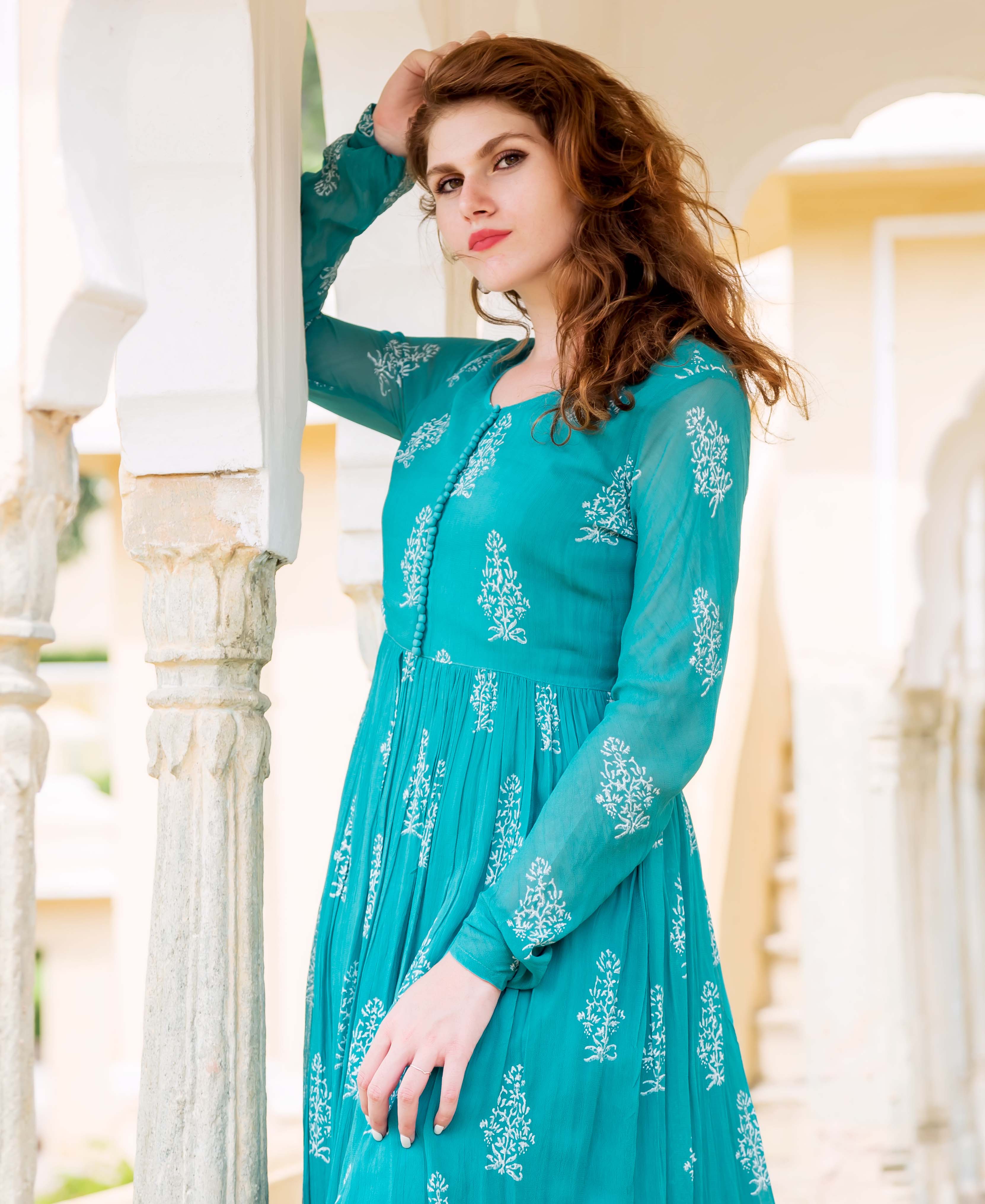 A woman in a teal dress with white block prints, posing beside a column.