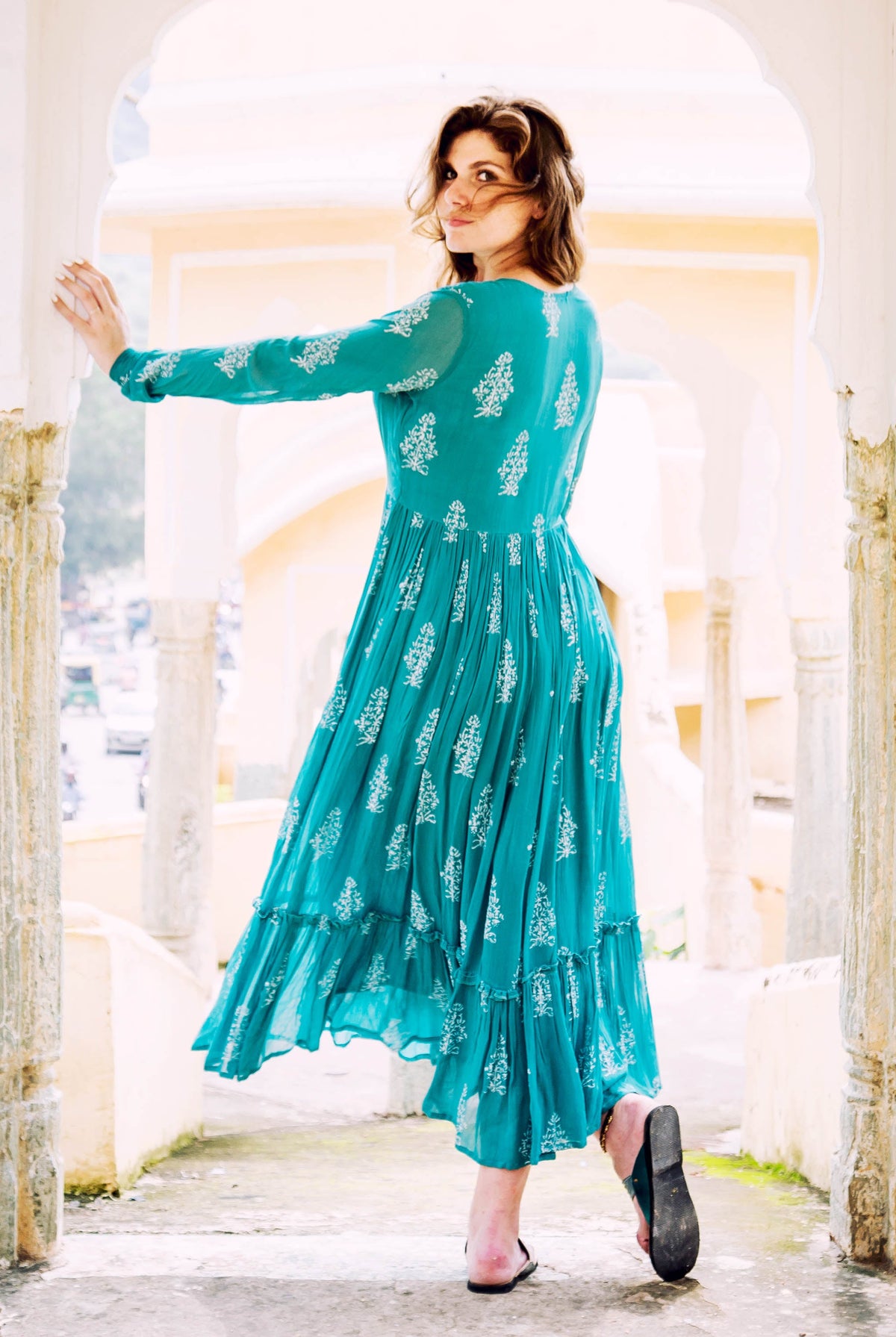A woman in a teal and white block-printed georgette dress, posing with her back to the camera.