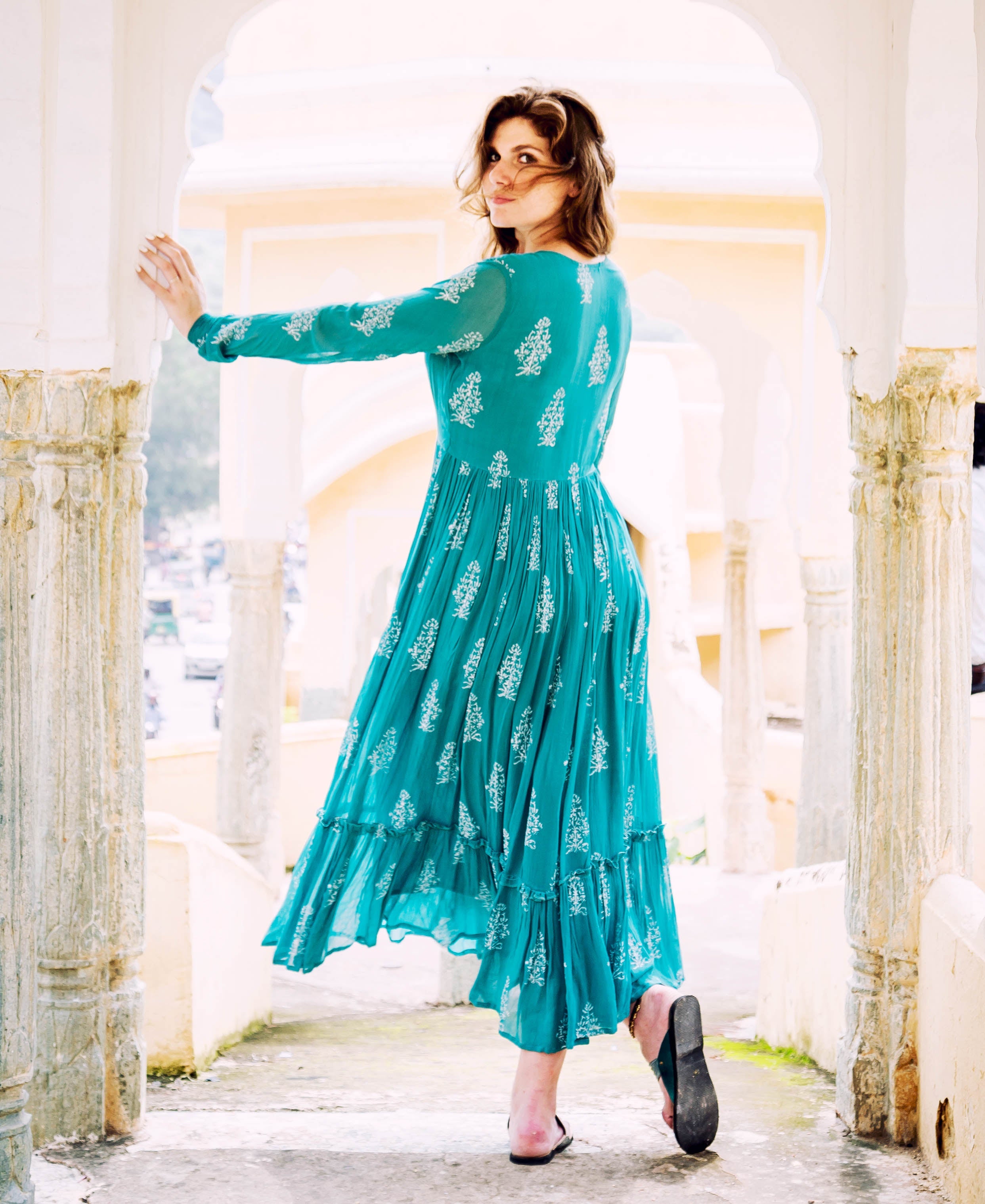 A woman in a teal and white block-printed georgette dress, posing with her back to the camera.