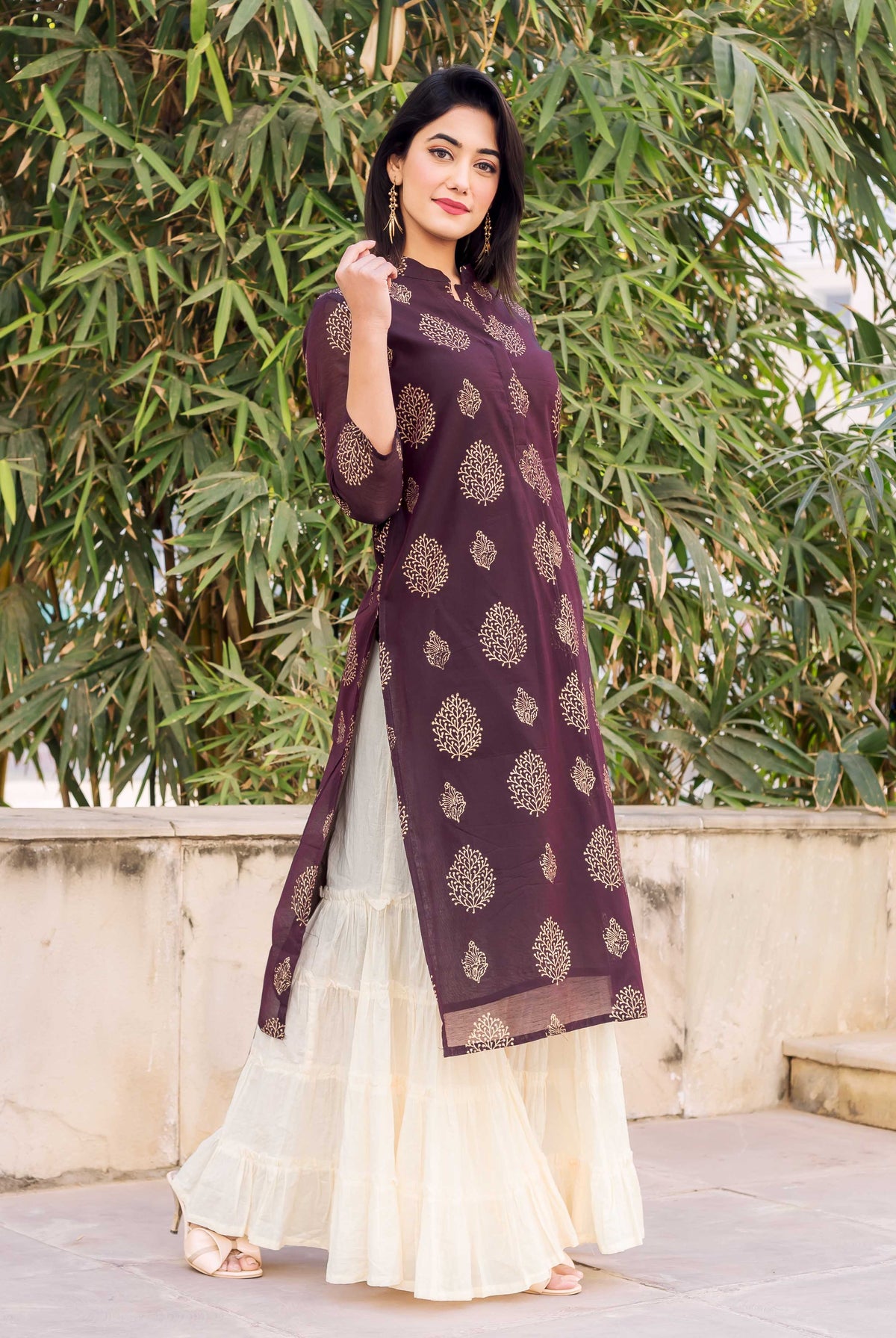 Model wearing a wine kurta with gold leaf print and a creme tiered sharara.