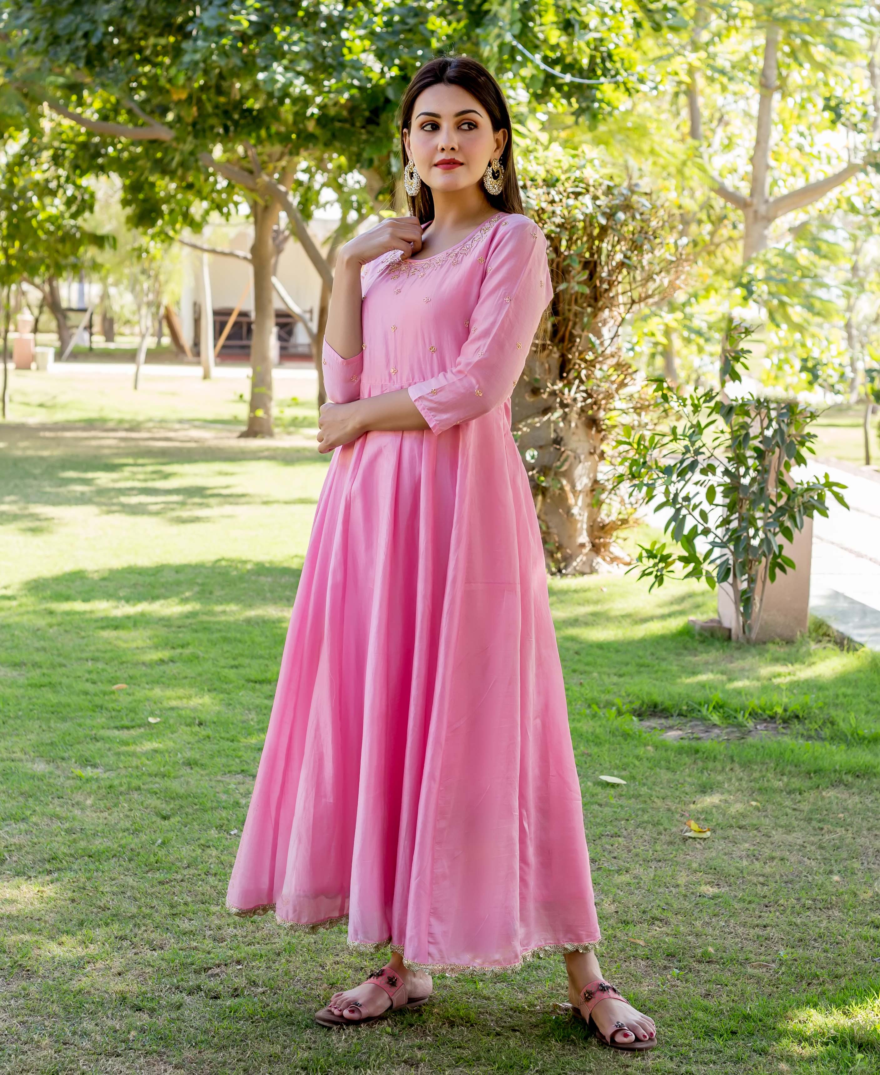 A model wearing a pink dress with gold embroidery, standing in a grassy area.