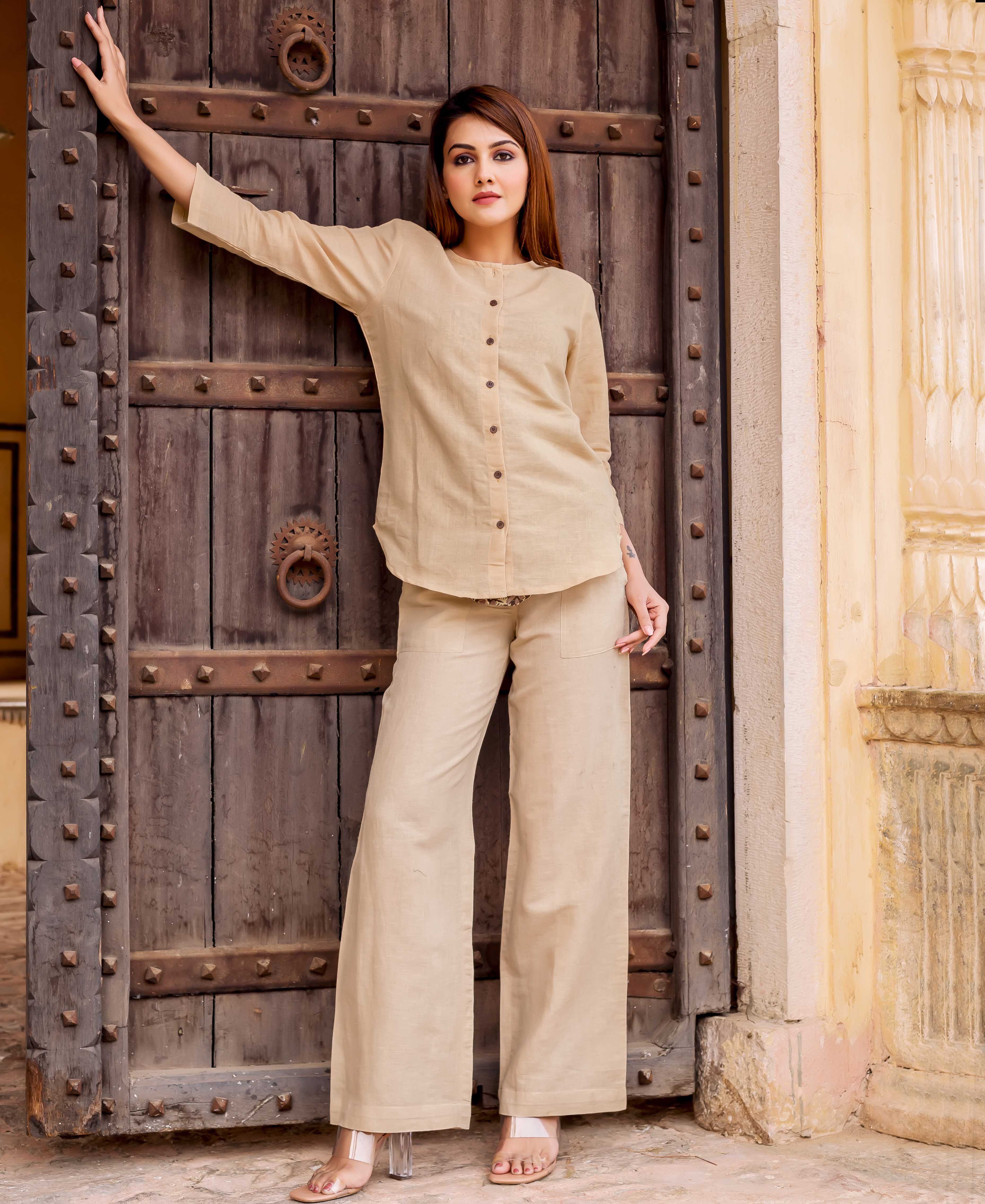 A woman in a beige linen coordinated set stands in front of a wooden door.