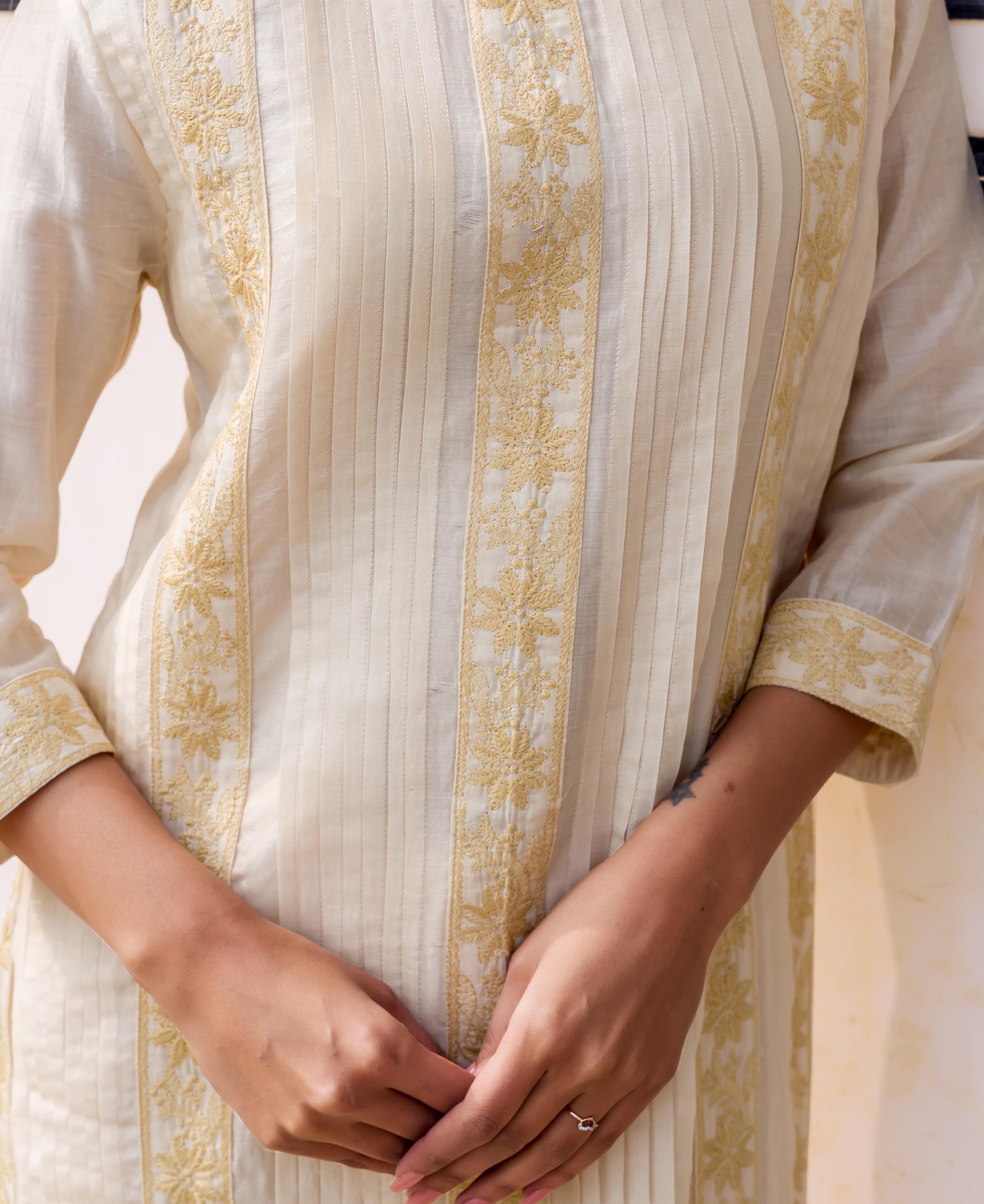 Model wearing off-white kurta featuring delicate golden floral embroidery and vertical pintucks.
