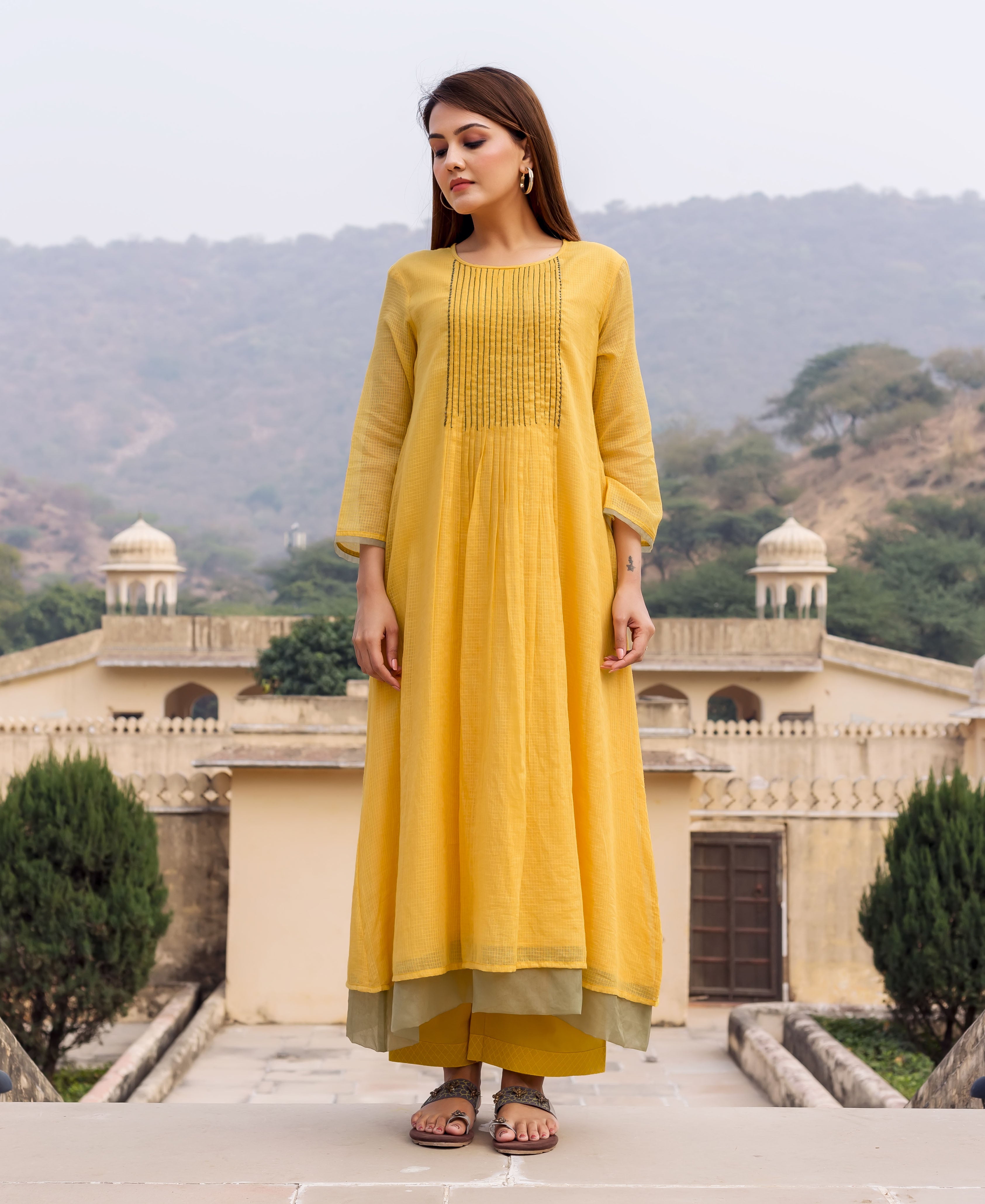 Front view of model wearing a honey yellow kurta, with embroidered pin-tucked yoke, and green layered pants.