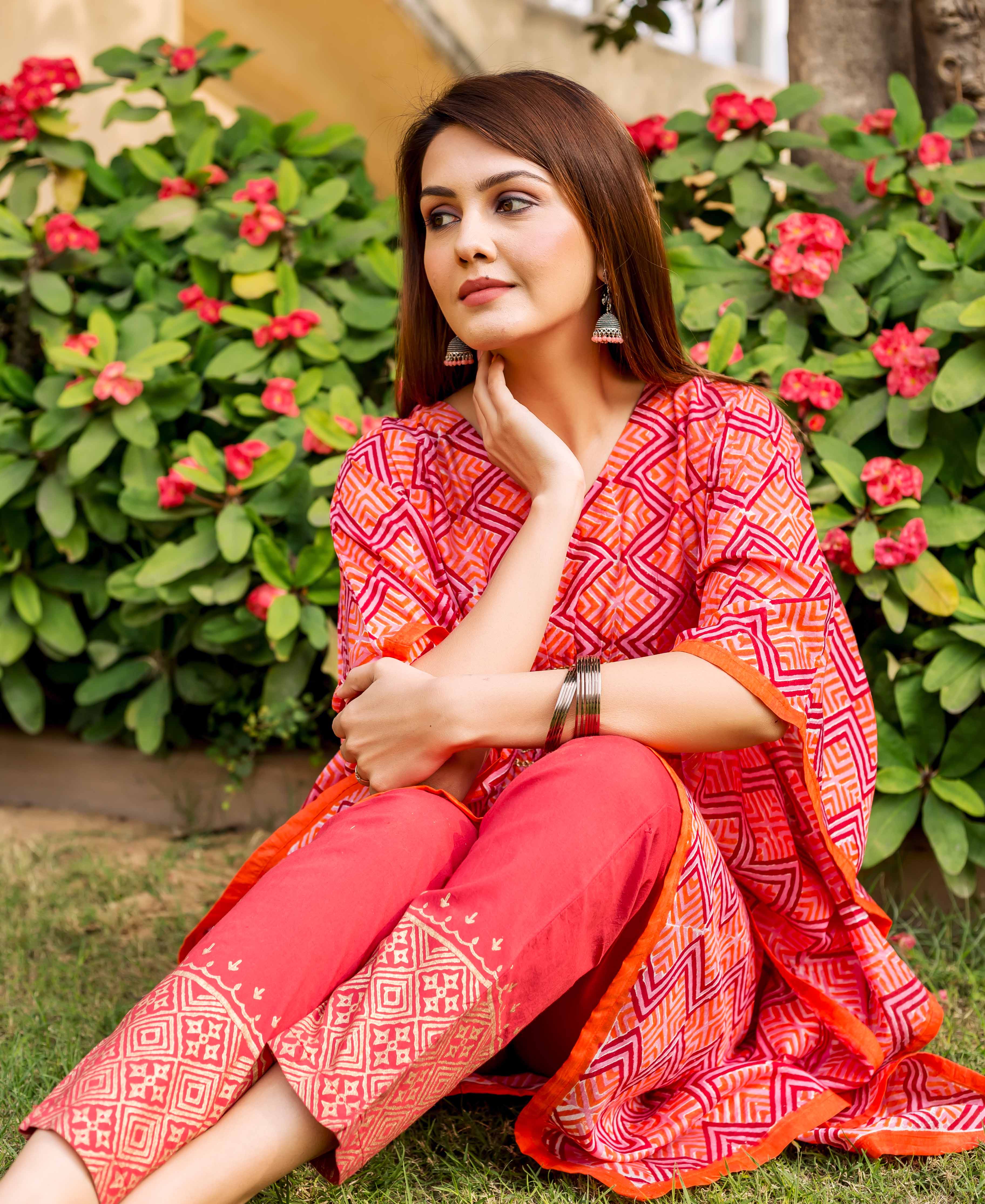 Model sitting on grass wearing a red-orange zig-zag printed kaftan and coral embroidered pants.