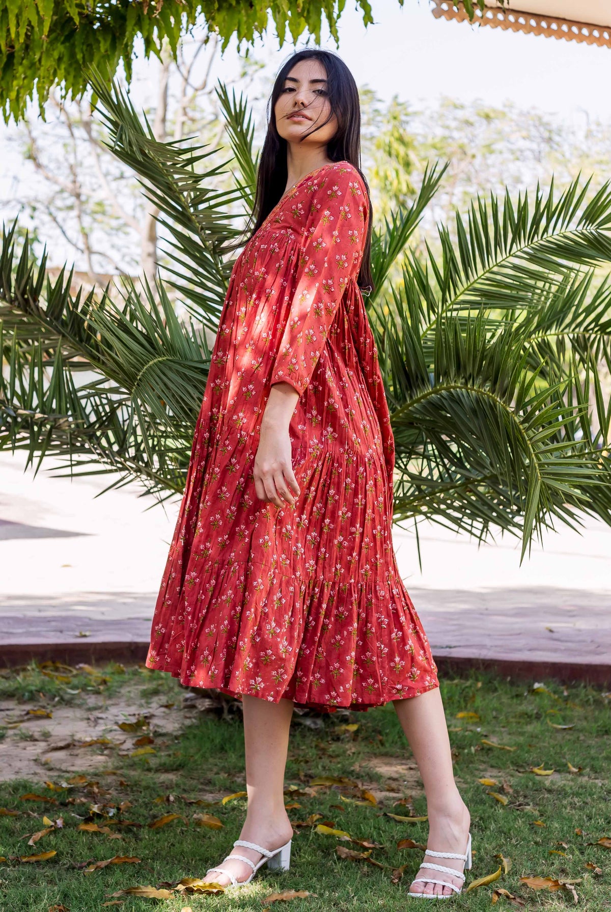 Model wears a coral-red tiered midi dress with small floral prints, outdoors.