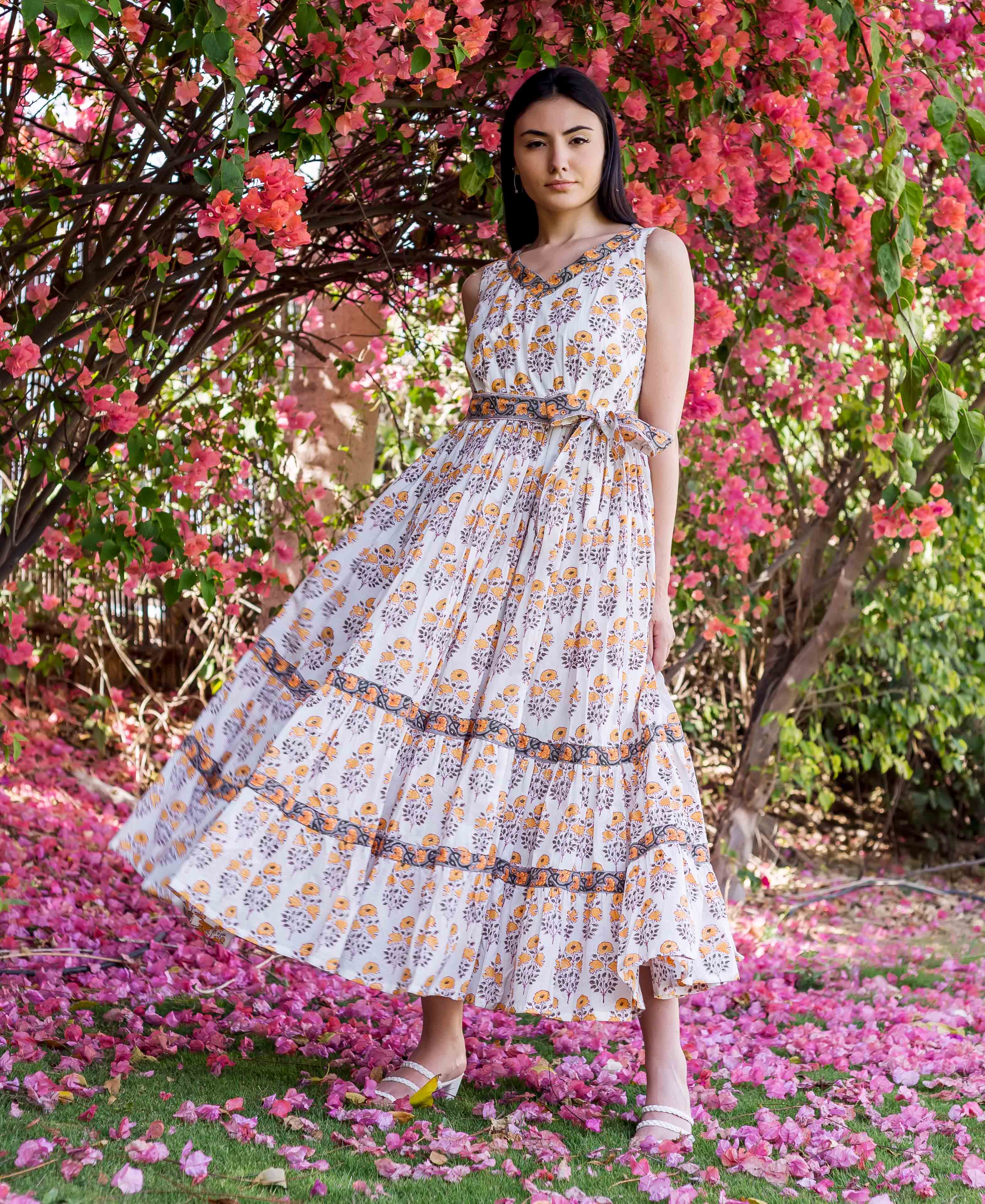 Full-body model wears white and yellow block printed tiered dress amidst vibrant pink flowers.