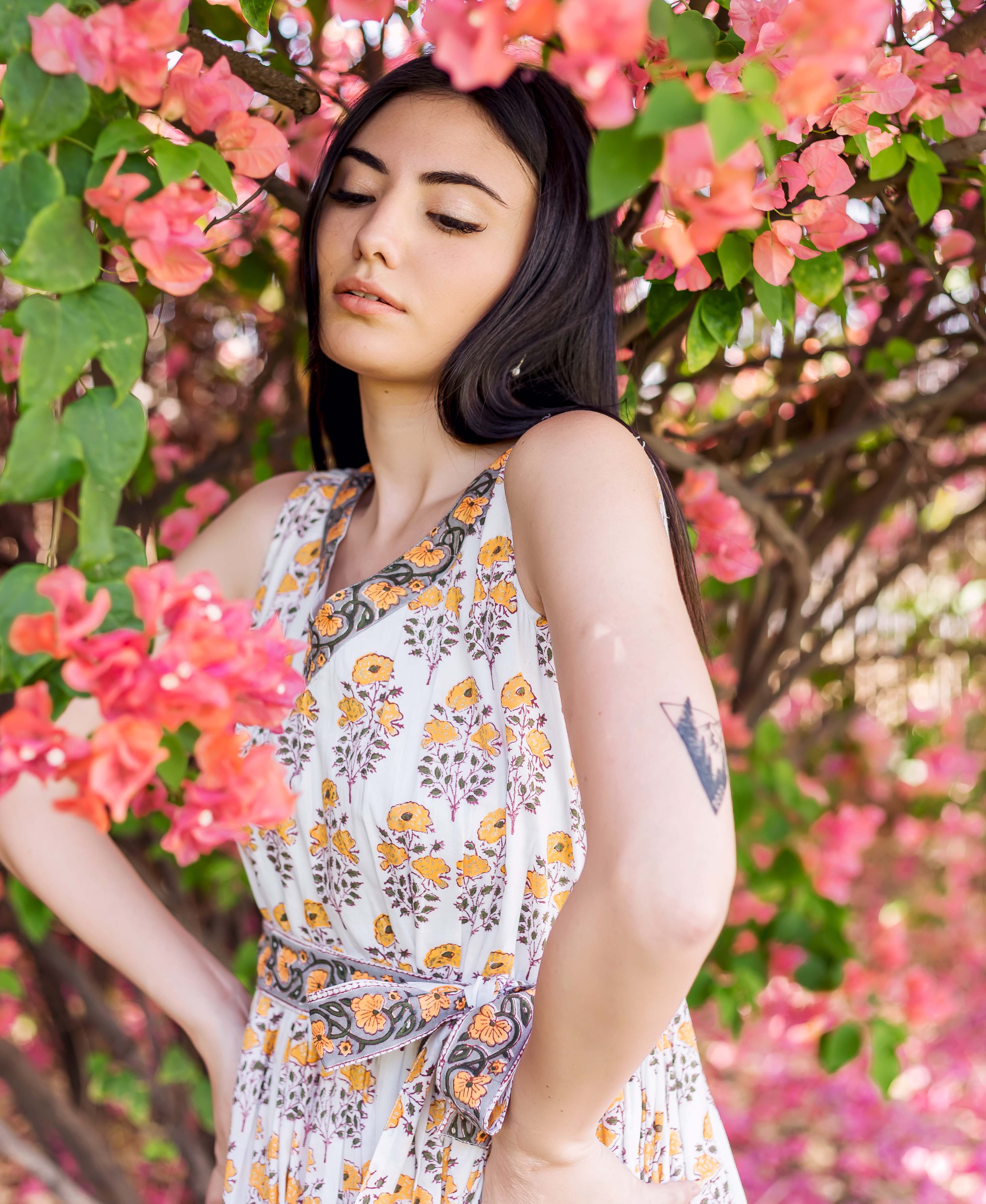 Model in white and yellow block-printed floral dress with waist tie, framed by pink flowers.
