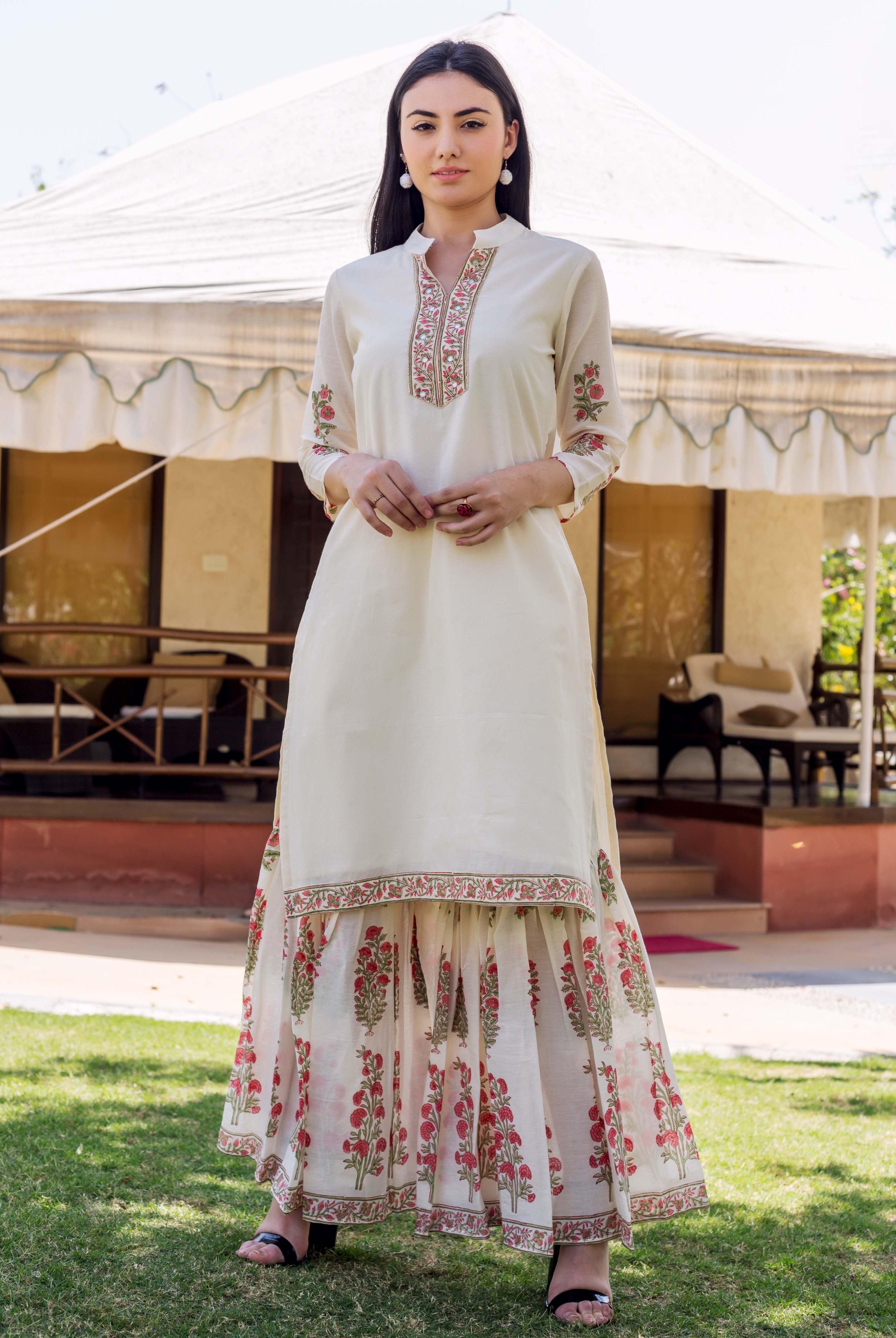 Model in ivory embroidered kurta and red floral block-printed tiered sharara on a lawn.