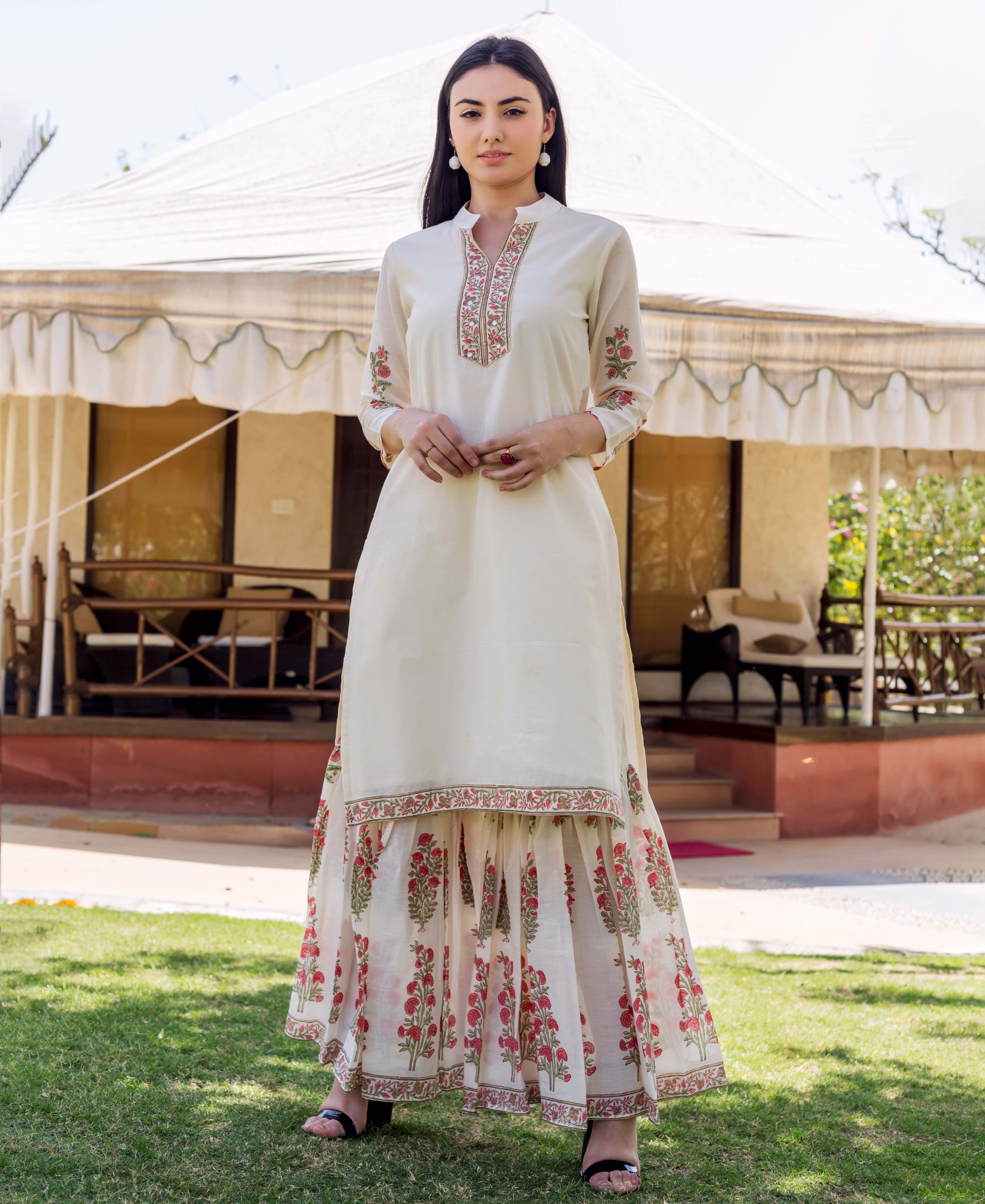 Model in ivory embroidered kurta and red floral block-printed tiered sharara on a lawn.