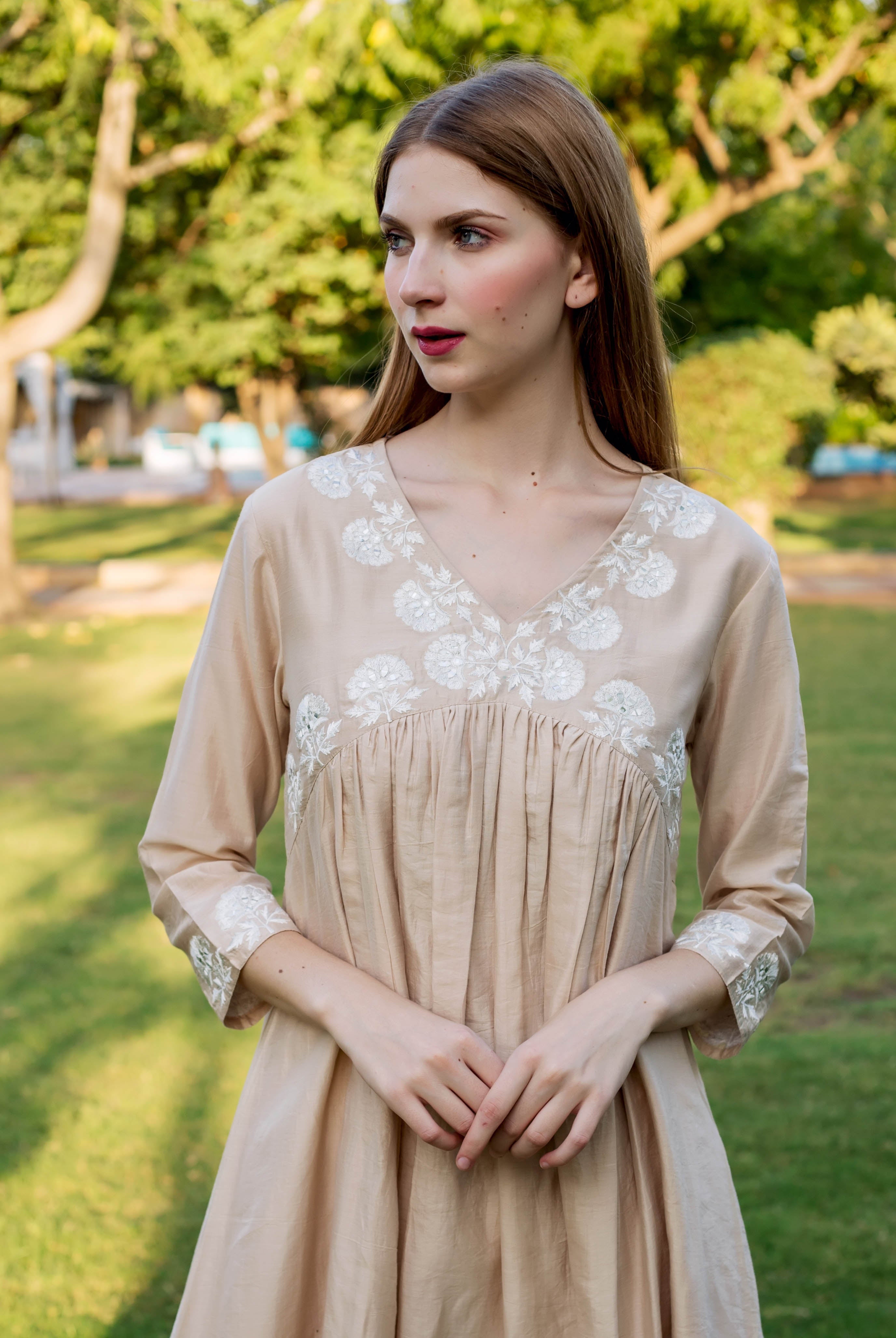 Beige dress with white embroidered floral details on bodice and sleeves, on a female model outdoors.
